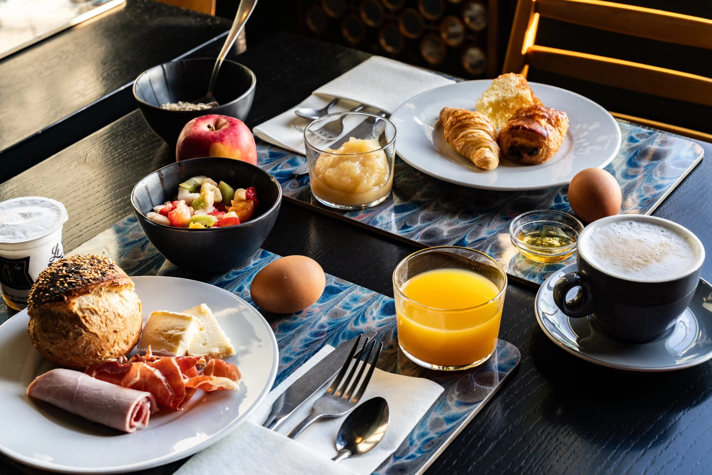 Petit déjeuner avec croissants, pain, œufs, jus d'orange, café, fruits, beurre, et miel sur une table en bois.