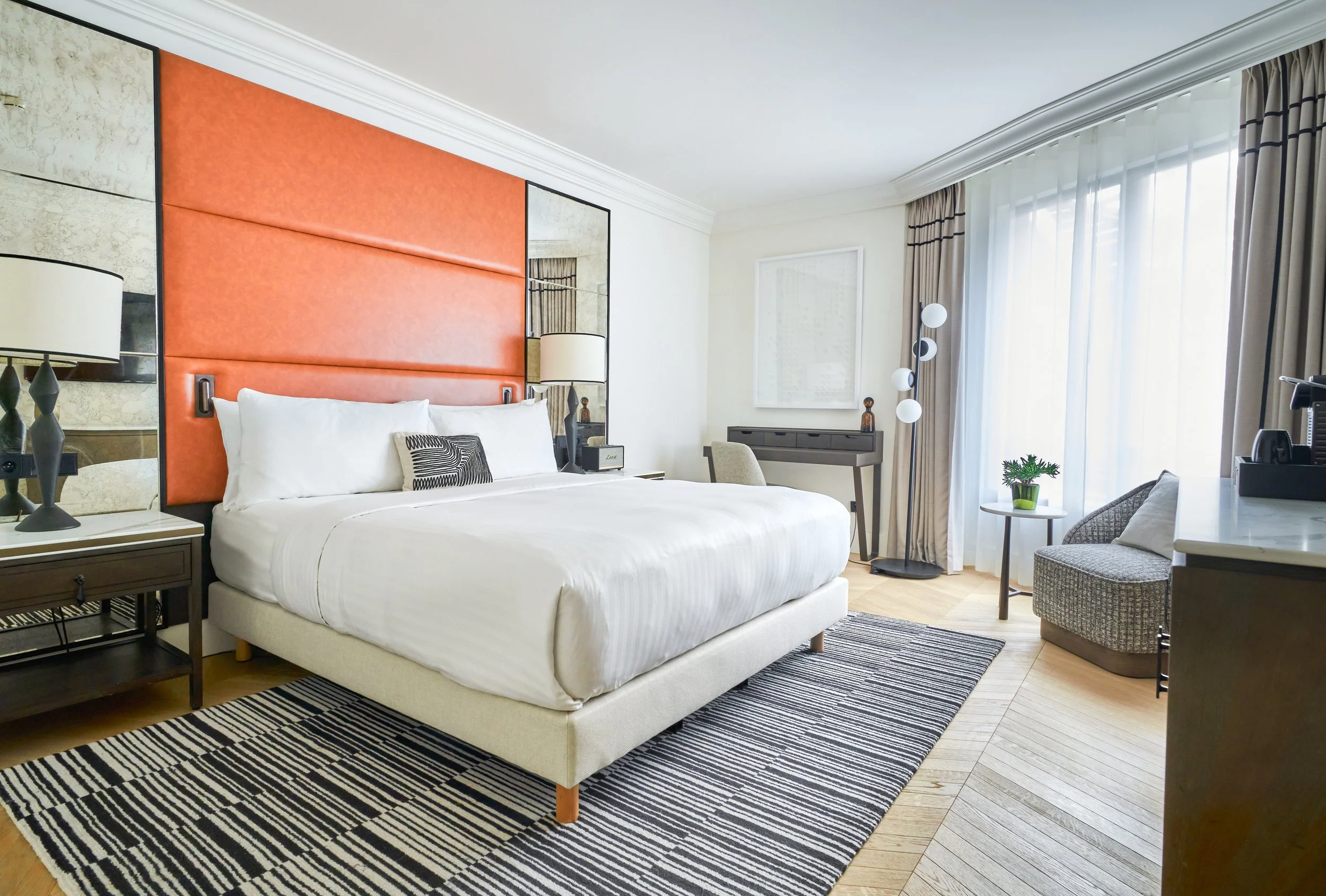 Chambre d'hôtel moderne avec un grand lit, tête de lit orange, lampes de chevet, tapis noir et blanc, petite table avec une plante, chaise, et une grande fenêtre avec des rideaux.