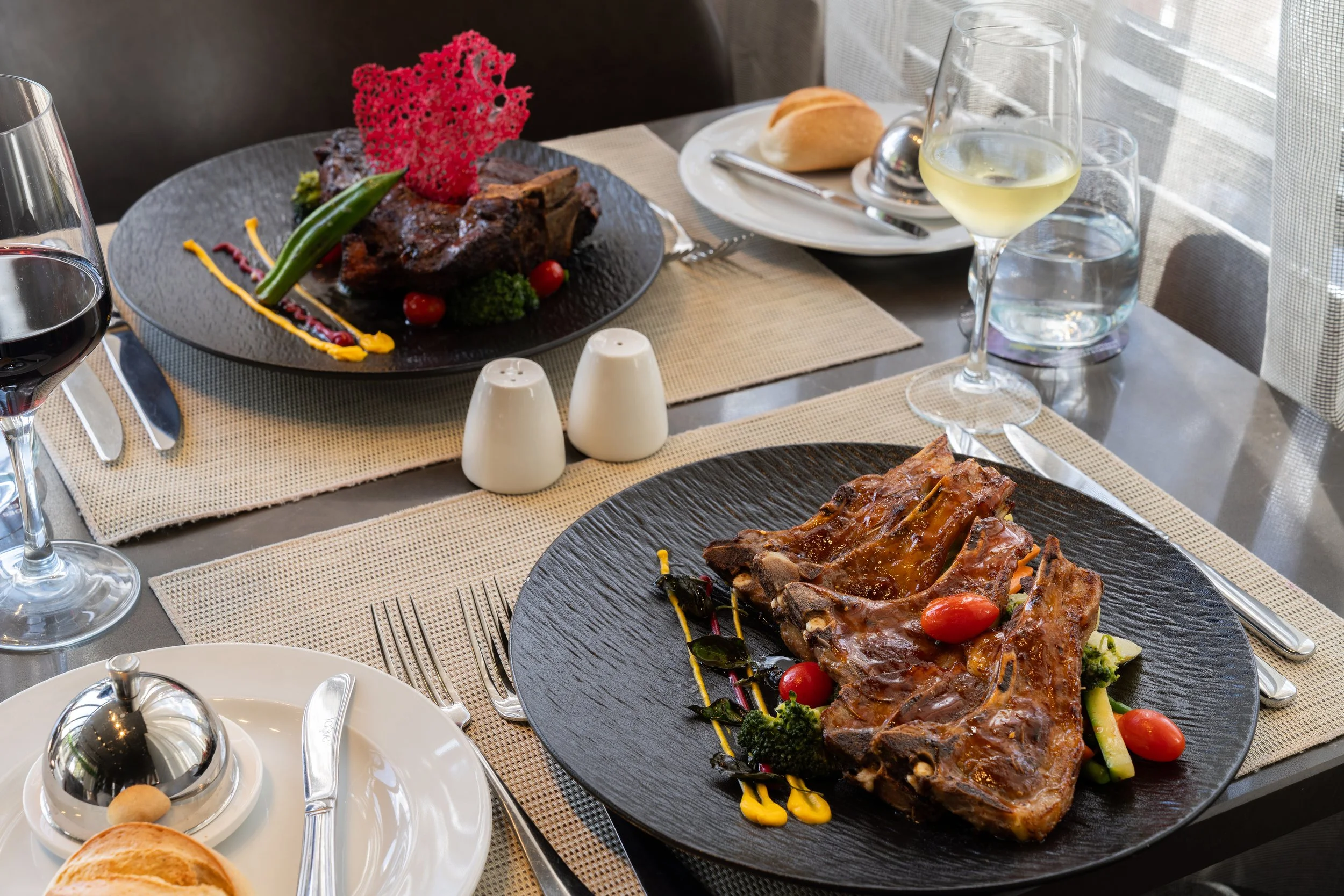 Deux plats principaux avec pièce de viande, légumes et sauces, accompagnés de verre de vin blanc et de verre d'eau, sur une table dressée pour un repas. Se trouvent également du pain et des assiettes avec des couverts.