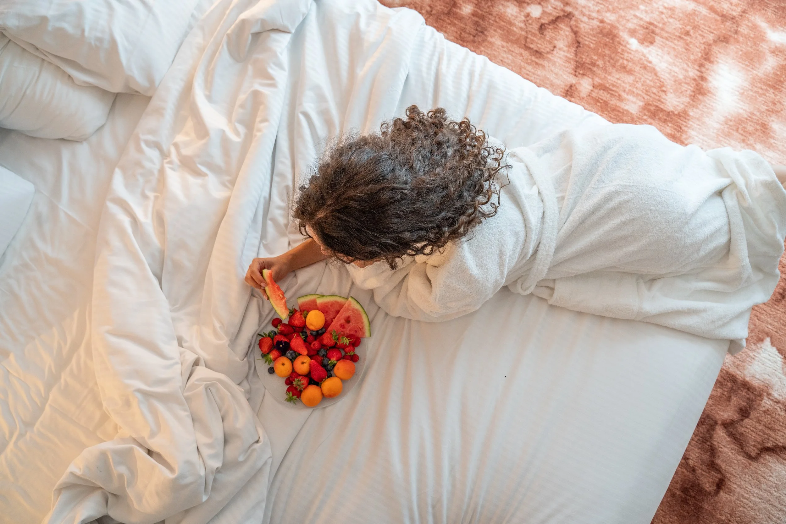 Femme en peignoir blanc mangeant un plateau de fruits sur un lit