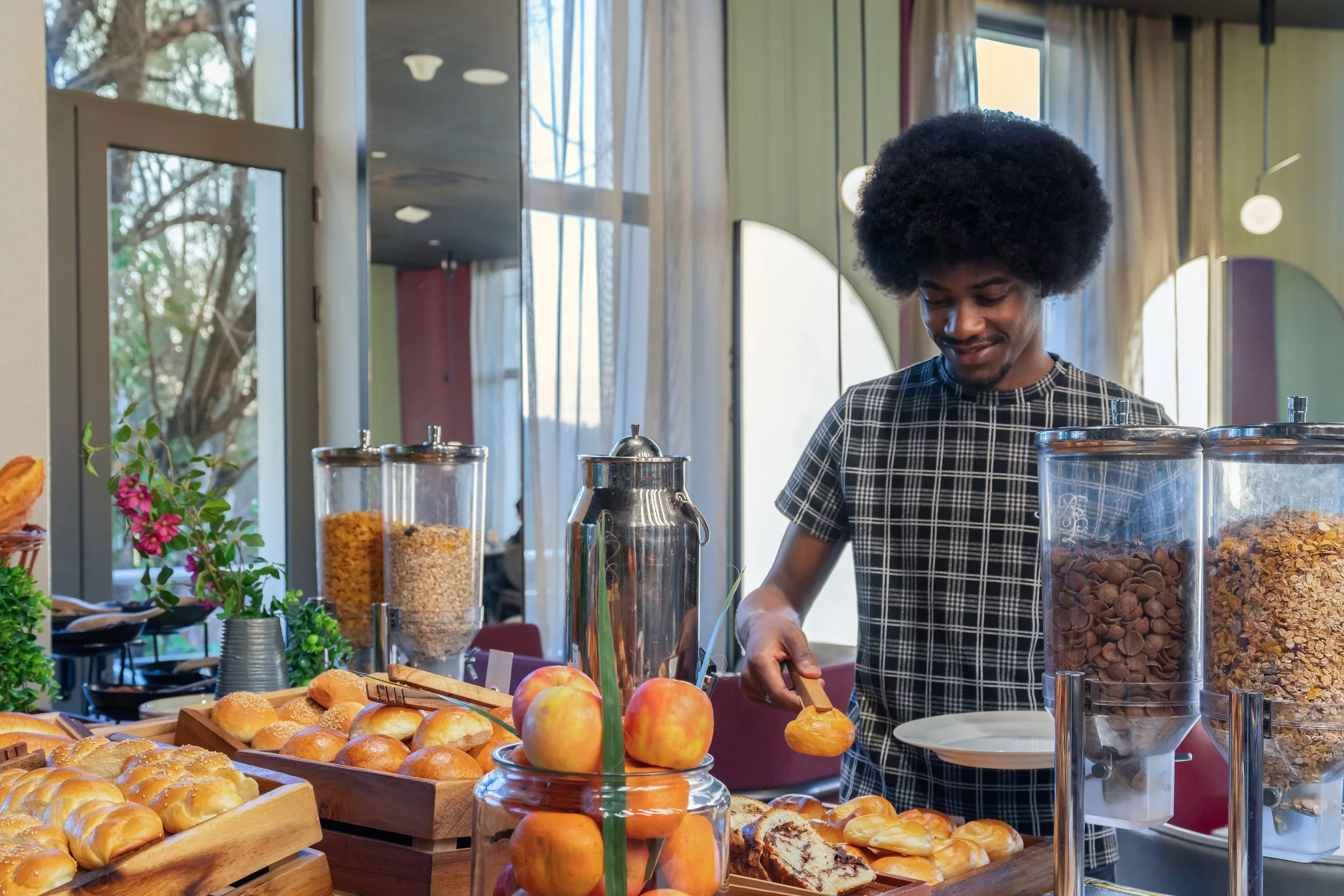 Un homme au visage souriant sert du café dans un buffet de petit-déjeuner avec des céréales, des fruits et des viennoiseries dans un restaurant lumineux.