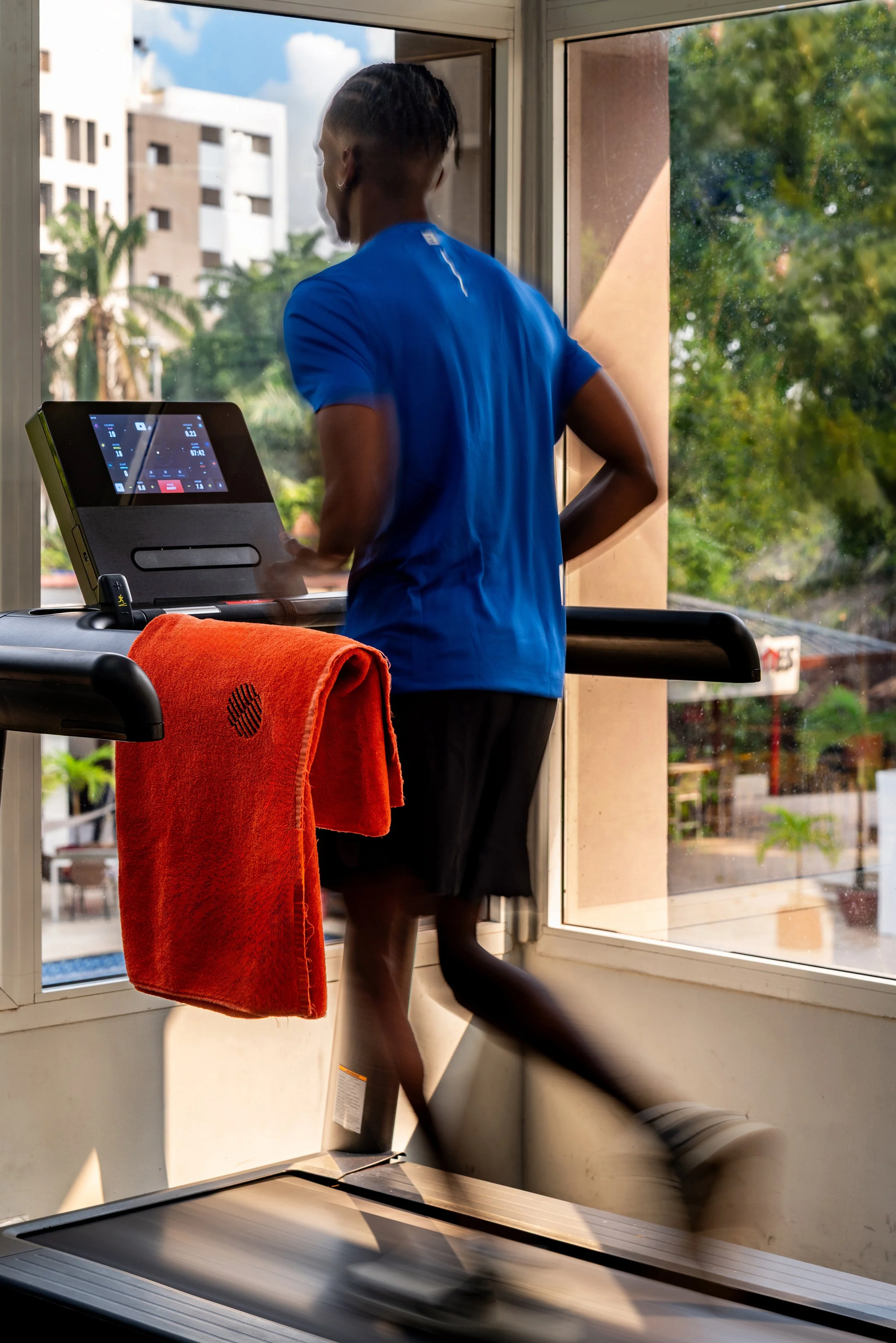 Personne en train de courir sur un tapis roulant dans une salle de sport, portant un t-shirt bleu et des shorts noirs, avec une serviette orange suspendue au guidon du tapis roulant, près d'une fenêtre offrant une vue sur des bâtiments et des arbres.