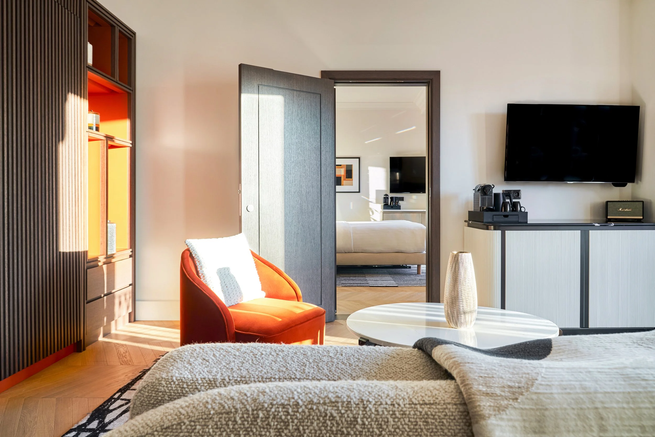 Salon lumineux de la chambre d'un hôtel de luxe, un fauteuil orange, une table basse blanche, un téléviseur à écran plat, et une baie vitrée laissant entrer la lumière naturelle.