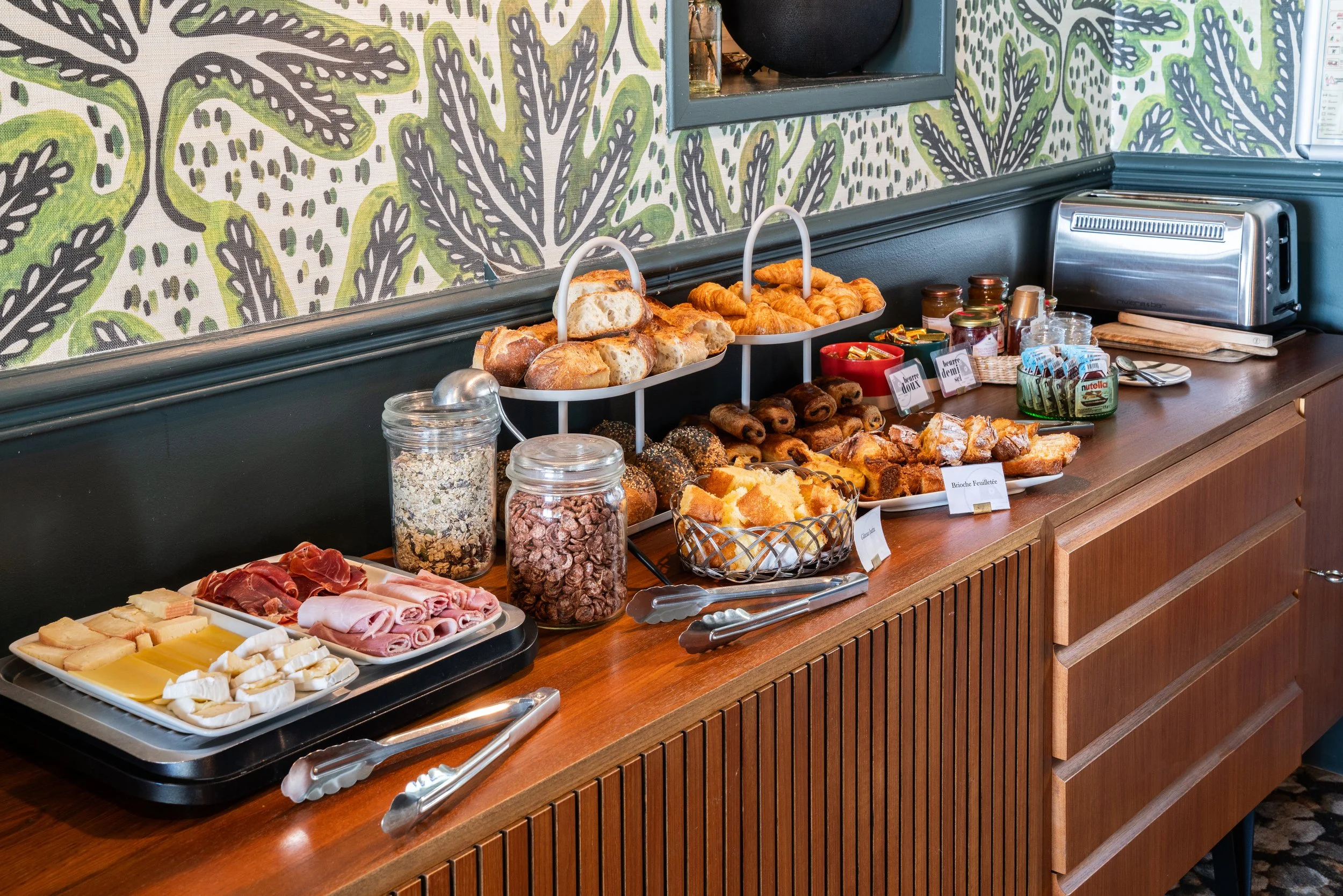 Buffet de petit-déjeuner avec diverses viennoiseries, pains, charcuterie, fromages, céréales, confitures sur un meuble en bois contre un mur décoratif.