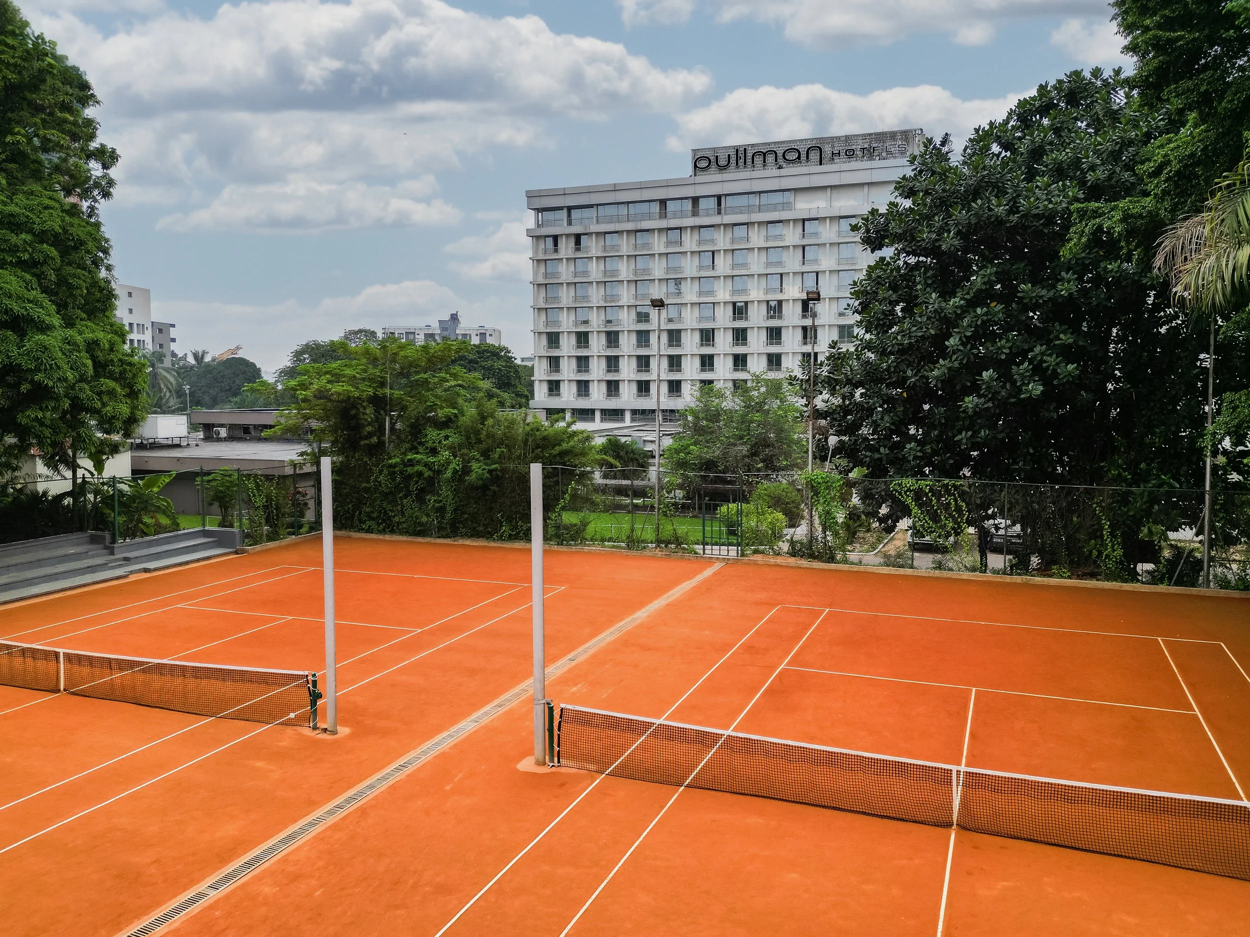 Court de tennis en terre battue orange, entouré d’arbres et d’un bâtiment haut avec le panneau 'Pullman Hotels' visible en arrière-plan.