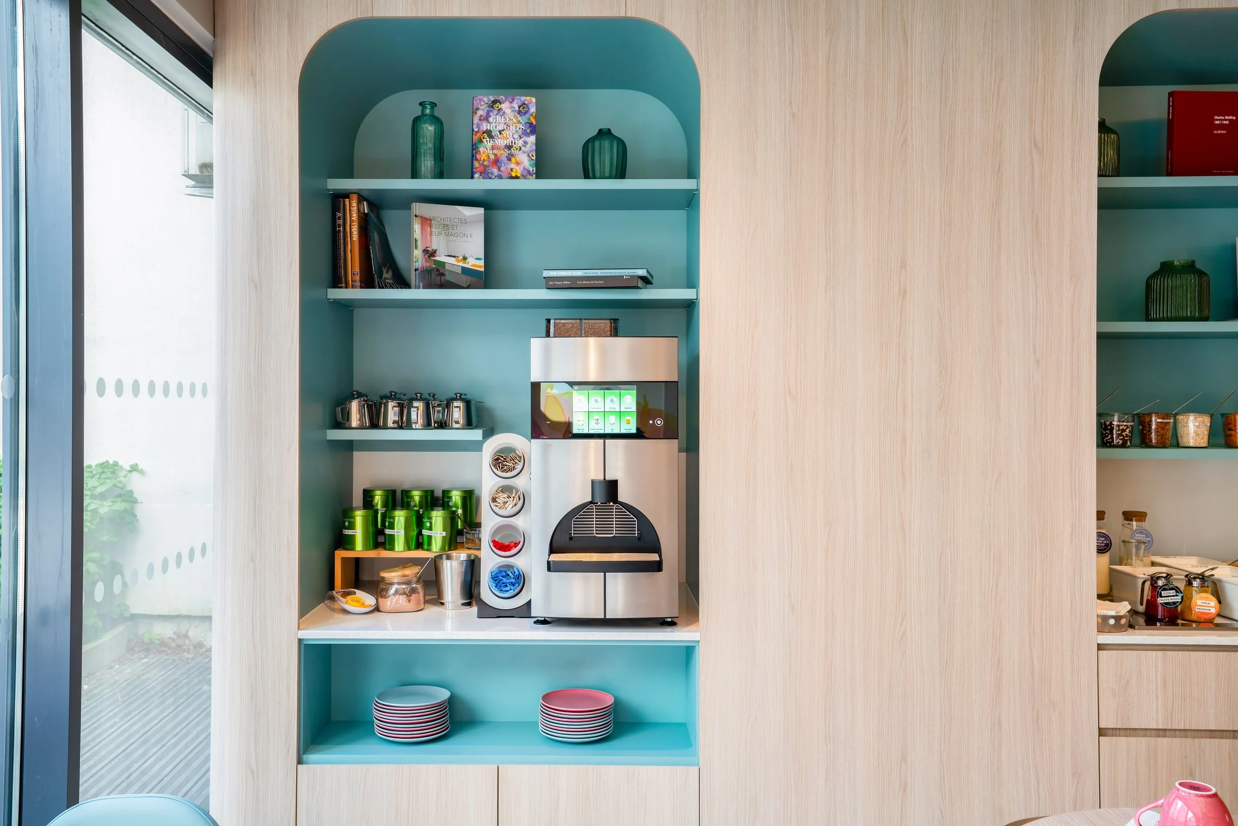Machine à café, étagères avec boites à thé, bocaux et livres, décor en bois clair et turquoise.