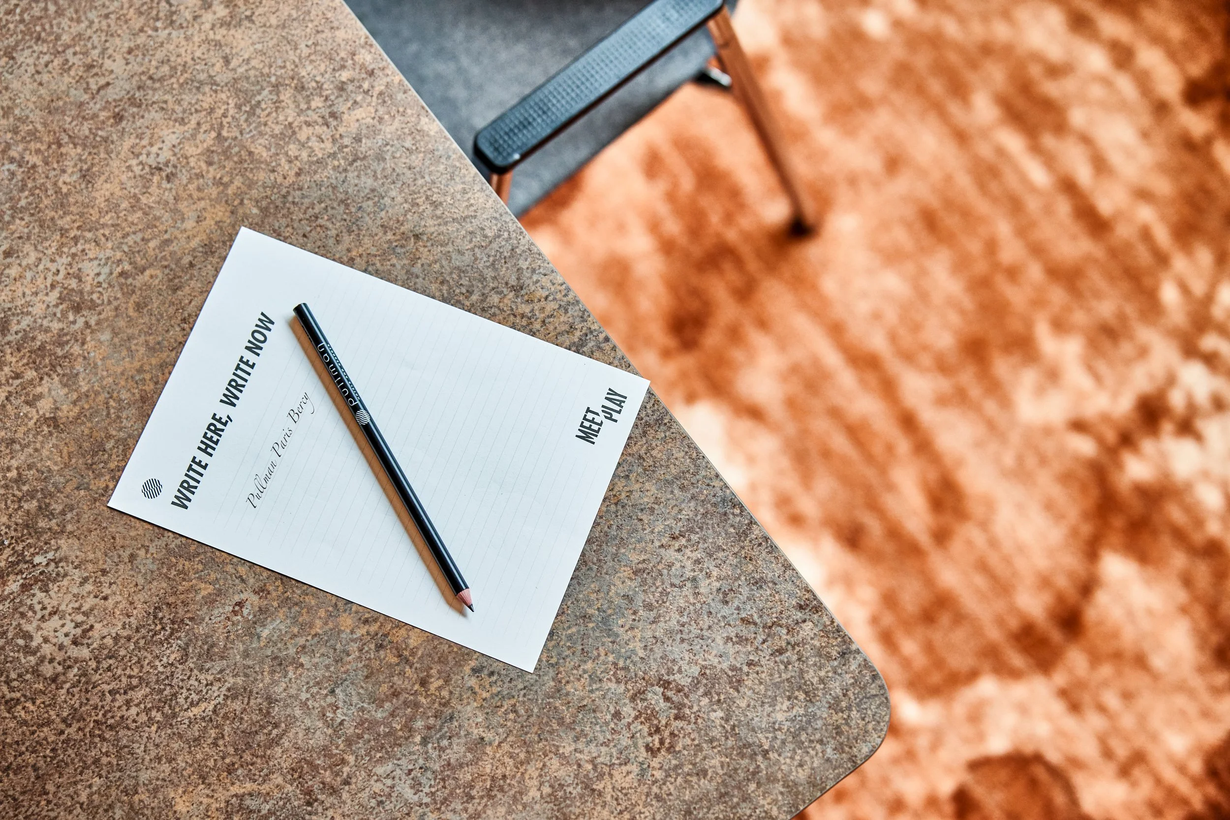 Un bloc note et un stylo noir posé dessus, sur une table en pierre, avec un fauteuil en arrière-plan sur un sol en terre cuite.