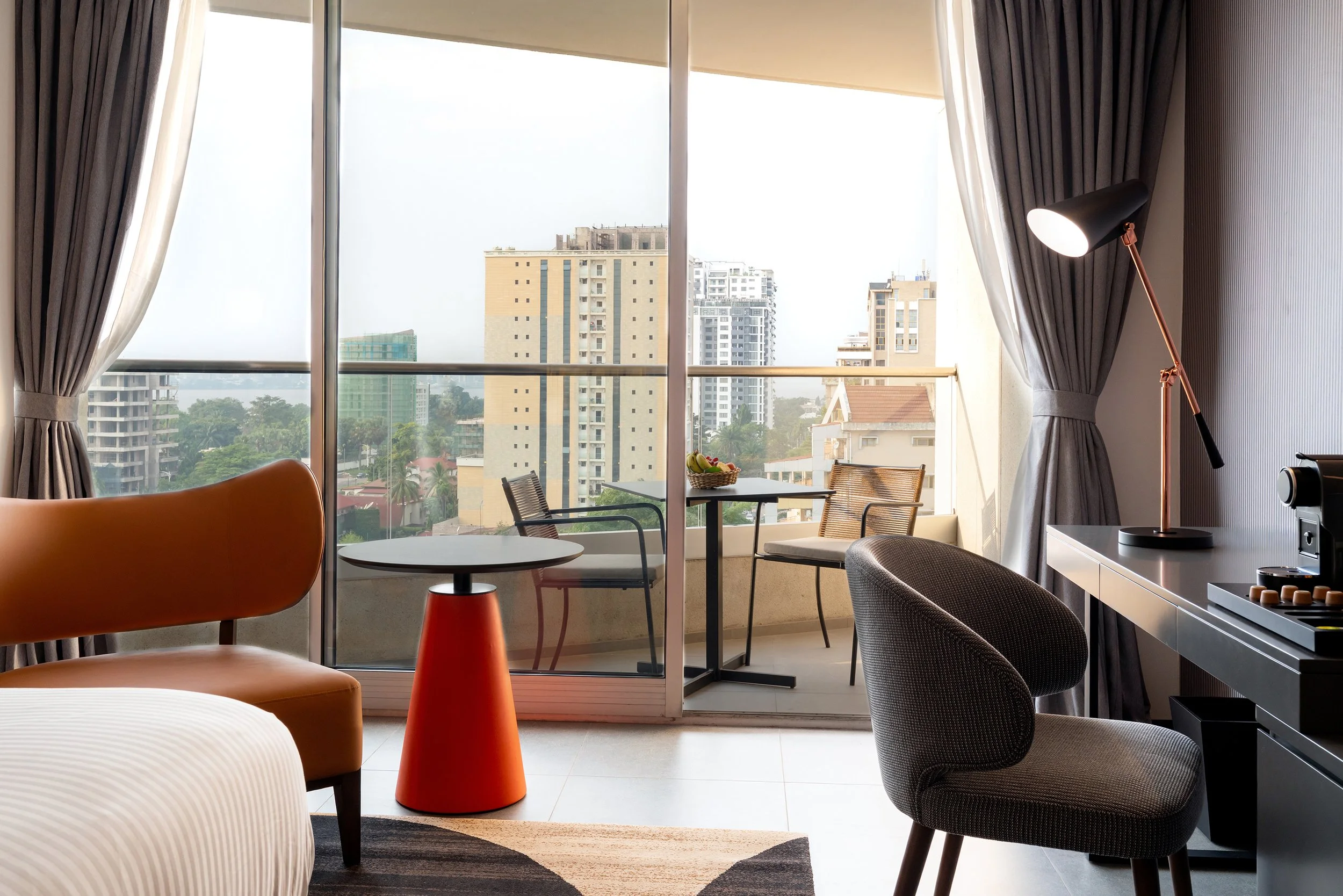 Salon d'une chambre d'hôtel avec un fauteuil orange, une table ronde, deux chaises sur un balcon, un bureau avec une lampe et une machine à café, grande baie vitrée avec vue sur des immeubles en ville.
