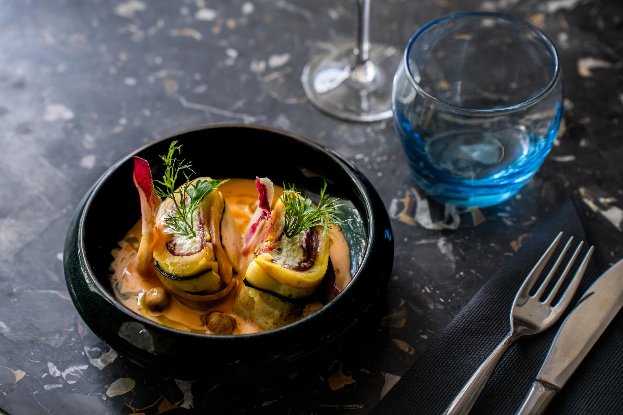 Assiette de roulés de courgettes garnis de viande, accompagnée de sauce crémeuse, décorée de fines herbes, avec un verre d'eau en verre bleu et un verre à vin vide, couverts en métal sur une serviette noire, pose sur une table en marbre