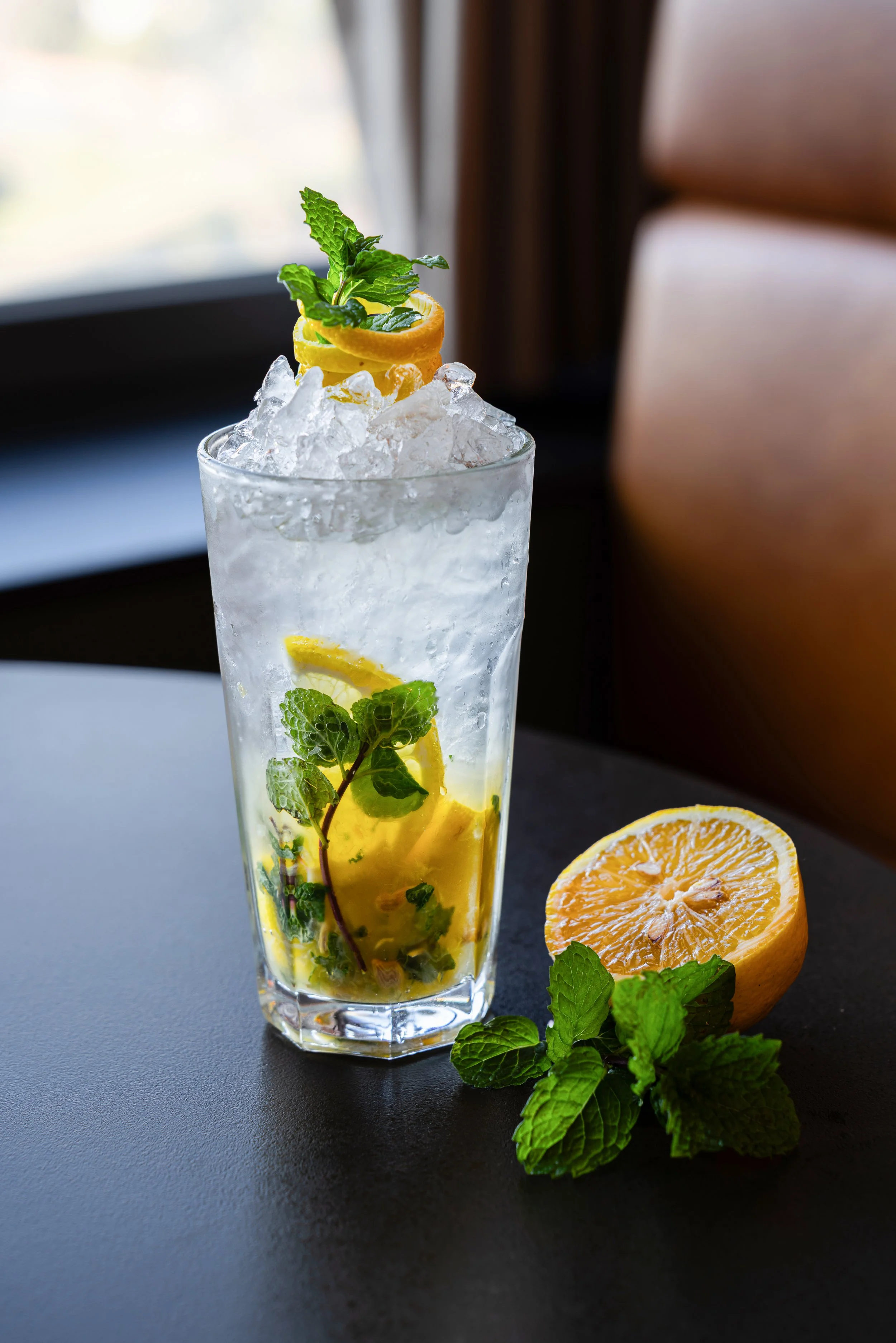 Cocktail avec glaçons, tranches de citron, rondelle de citron sur le dessus, feuilles de menthe, et une moitié de citron posé à côté avec des feuilles de menthe.