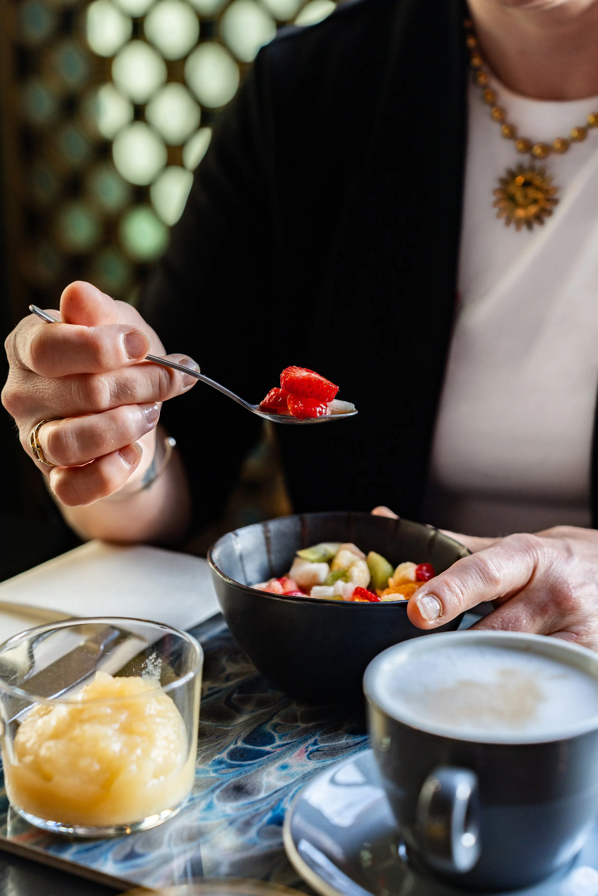 Une personne tient une cuillère avec des fraises coupées, un bol de salade de fruits est devant, ainsi qu'une tasse de café et un verre de sorbet ou de glace à gauche.