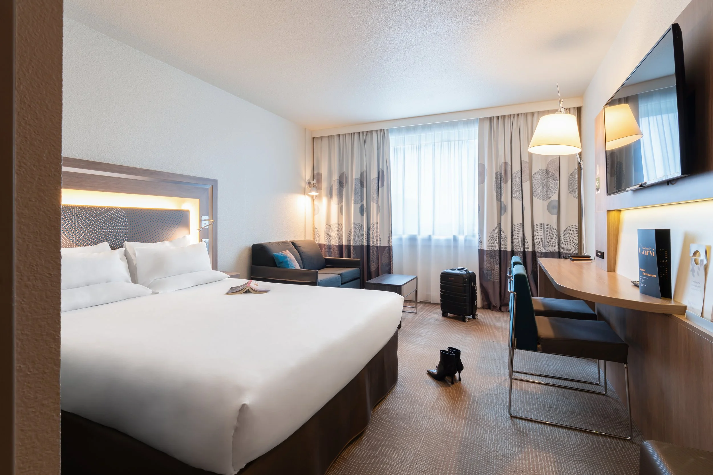 Chambre d'hôtel avec lit, fauteuils, télévision, bureau et valise.
