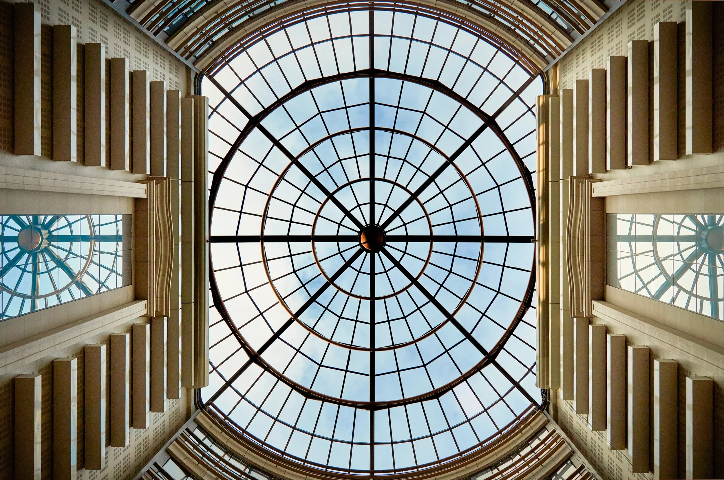 Vue intérieure d'un plafond en verre circulaire avec une structure en métal, entourée par des murs avec des fenêtres rectangulaires.