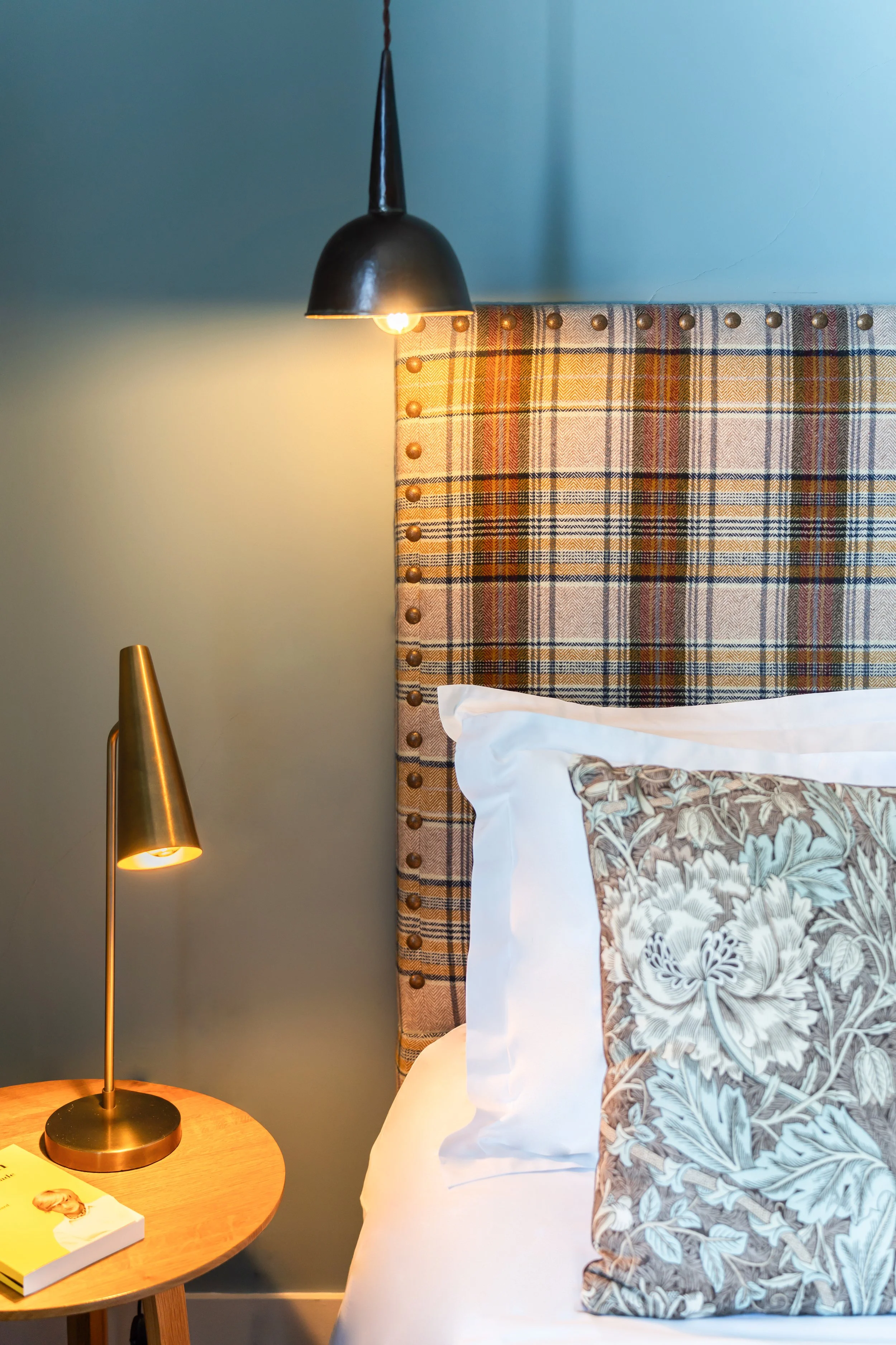 Coin de lit avec coussin floral, tête de lit à carreaux, lampe suspendue noire et lampe de table dorée, lampe de chevet en bois avec un livre dessus dans une chambre