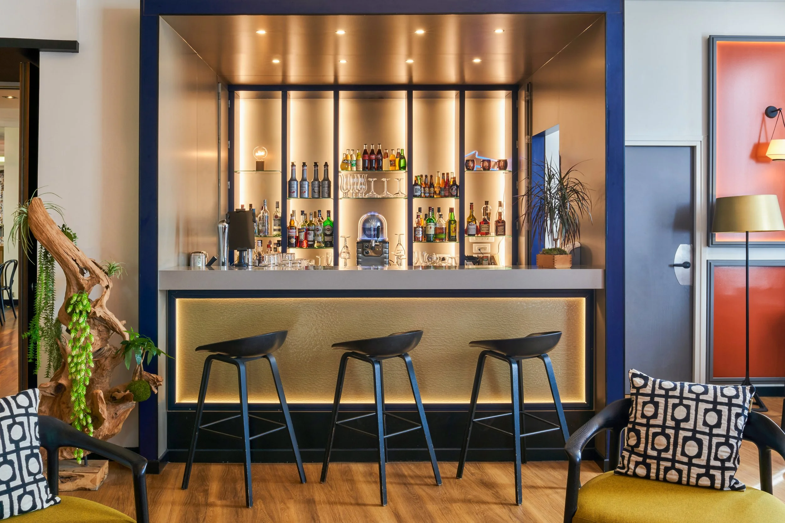 Un bar moderne avec un comptoir en façade finie en cuir synthétique beige, trois tabourets noirs en face, derrière un mur avec étagères lumineuses contenant des bouteilles d'alcool et des verres, décoration avec plantes et coussins à motifs