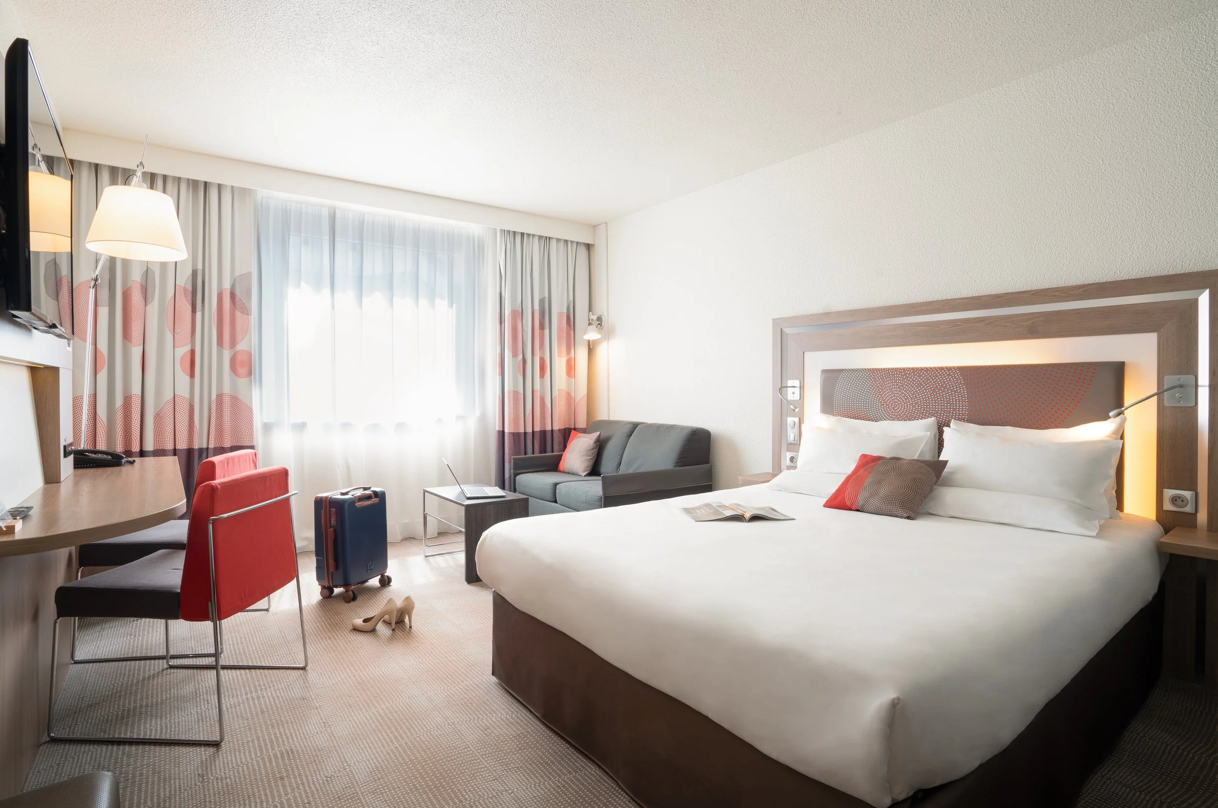 Chambre d'hôtel avec un lit, un canapé, une télévision, une valise, une paire de chaussures, un bureau avec deux chaises, des rideaux à motifs modernes et une lampe