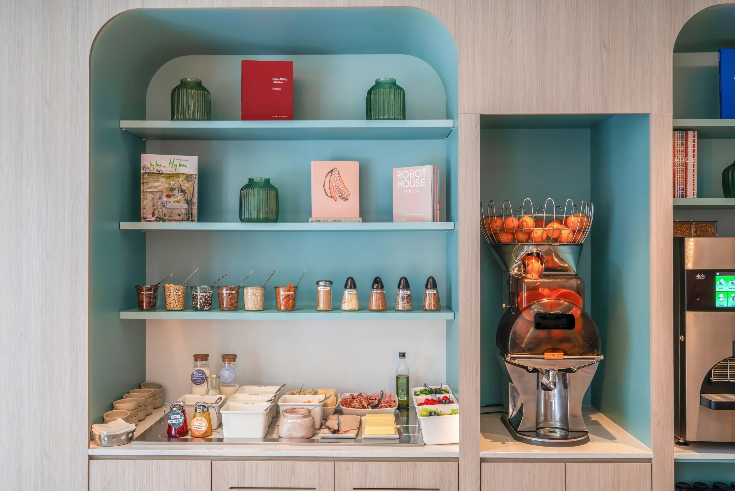 Un buffet de petit déjeuner avec un distributeur de fruits, plusieurs bocaux avec des céréales, un plateau avec des tranches de charcuterie, des œufs, du beurre, du fromage, des confitures et des sauces, ainsi qu'un espace avec des livres et des vase