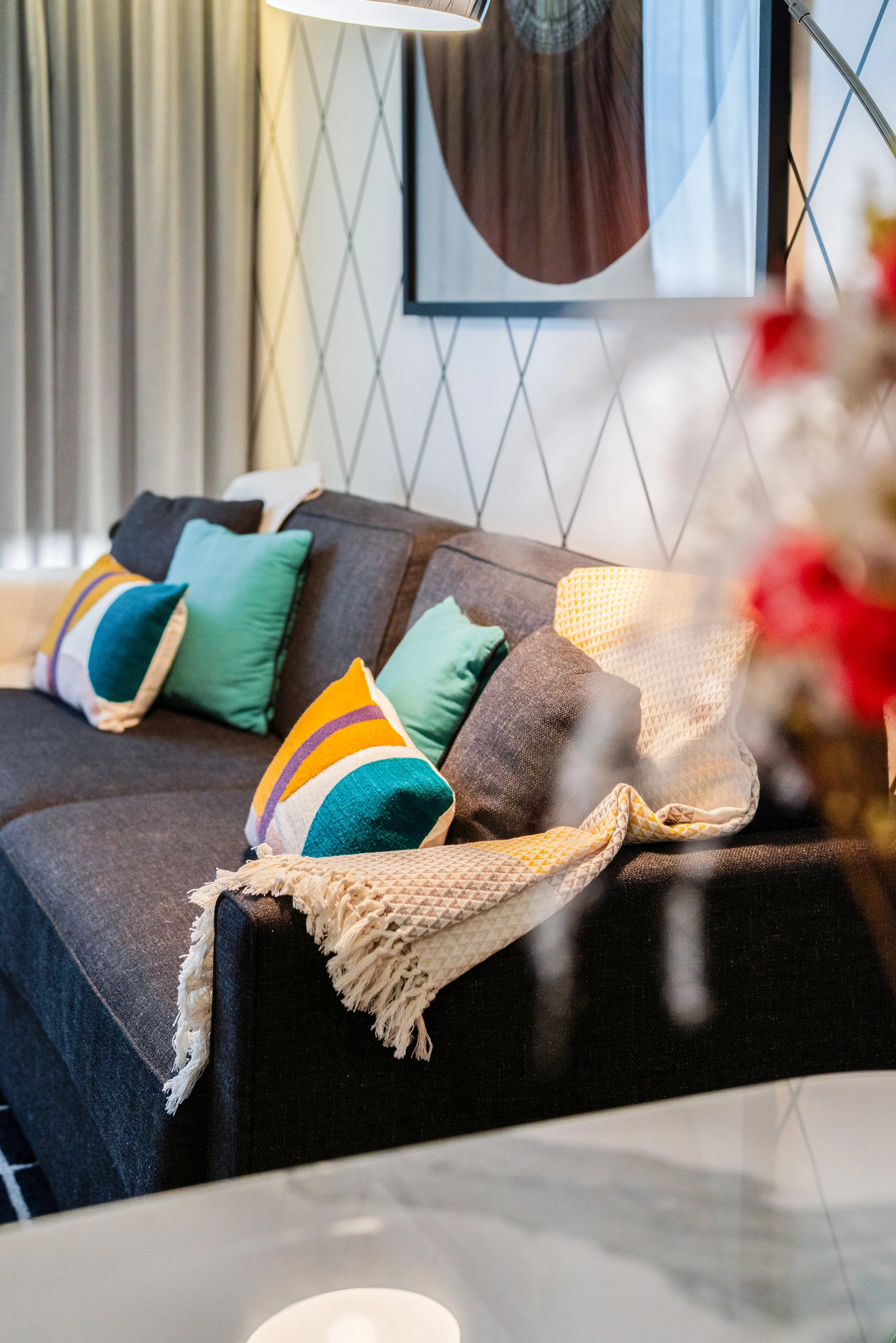 Salon moderne avec un canapé gris décoré avec des coussins colorés et un plaid blanc, mur avec motif géométrique, lampe de lecture et tableau abstrait.