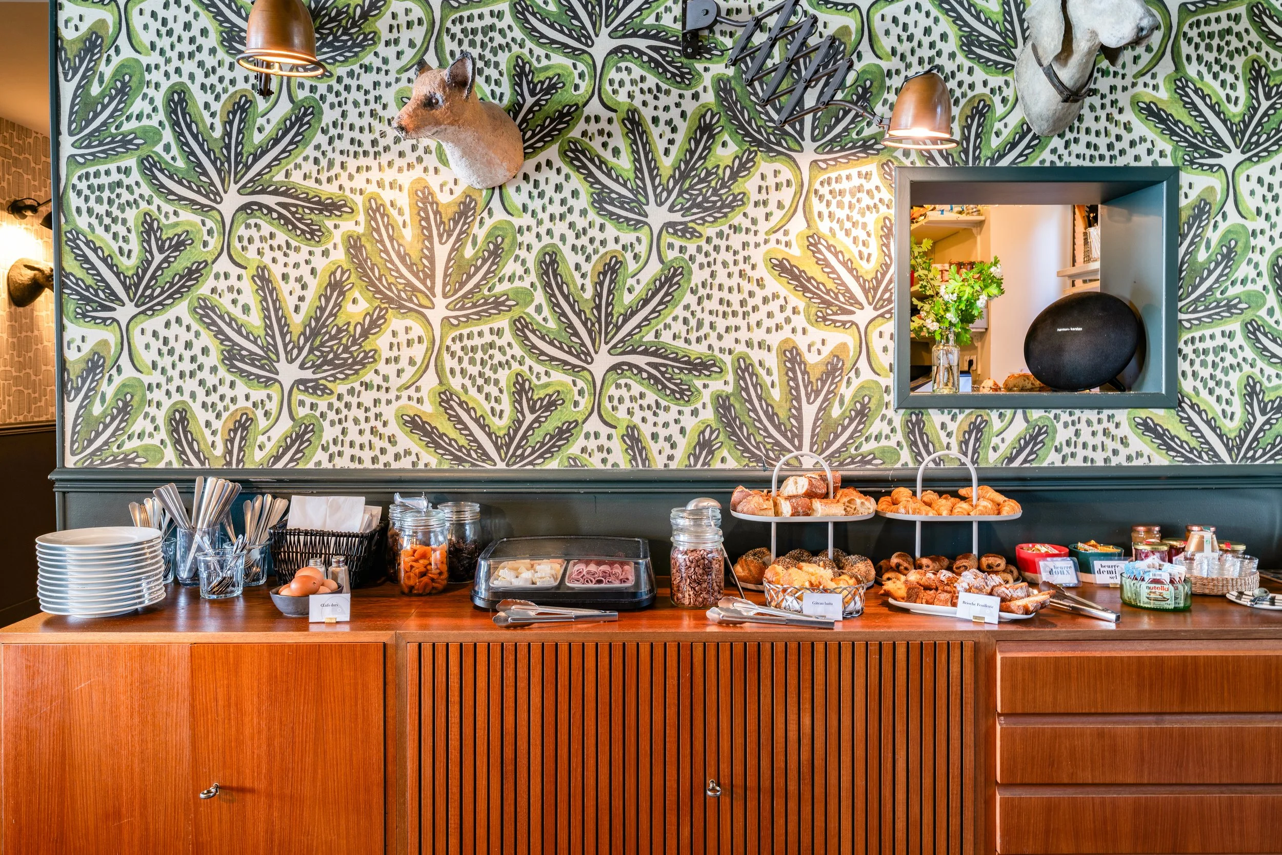 Buffet de petit déjeuner avec variétés de pâtisseries, fromages, confitures, œufs, et autres condiments, contre un mur décoré de papier peint à motif vert et noir avec des trophées d'animaux