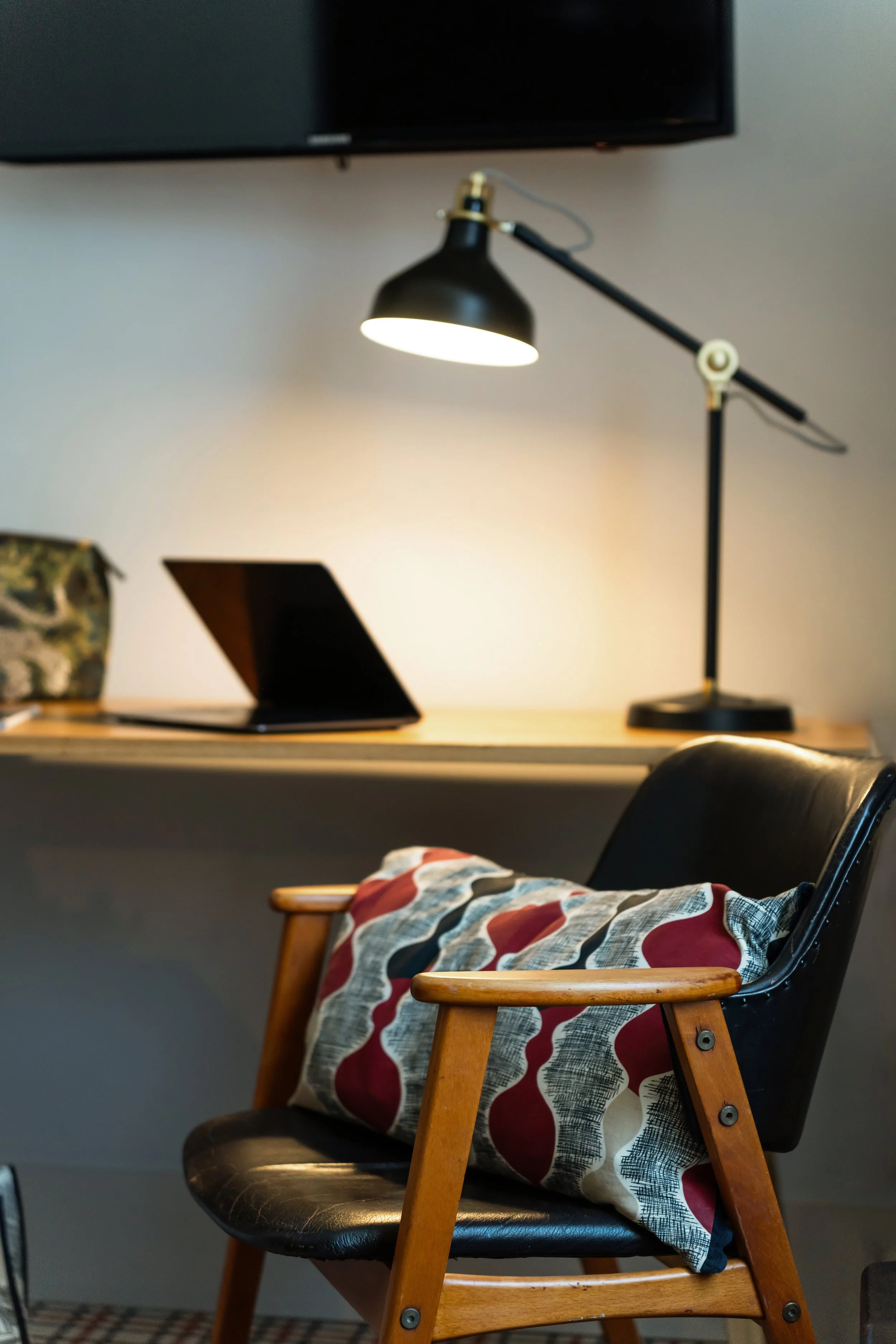 Un fauteuil en bois avec un coussin à motifs rouges et gris, placé devant un bureau avec une lampe de bureau noire inclinée et un ordinateur portable ouvert, dans un espace de travail moderne.