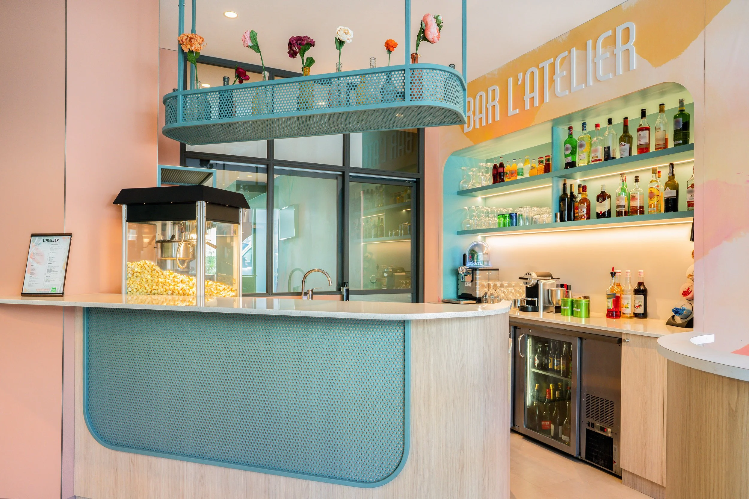 Un bar coloré avec un comptoir en formica, des étagères avec des bouteilles d'alcool, un distributeur de popcorn, et une décoration tendance.