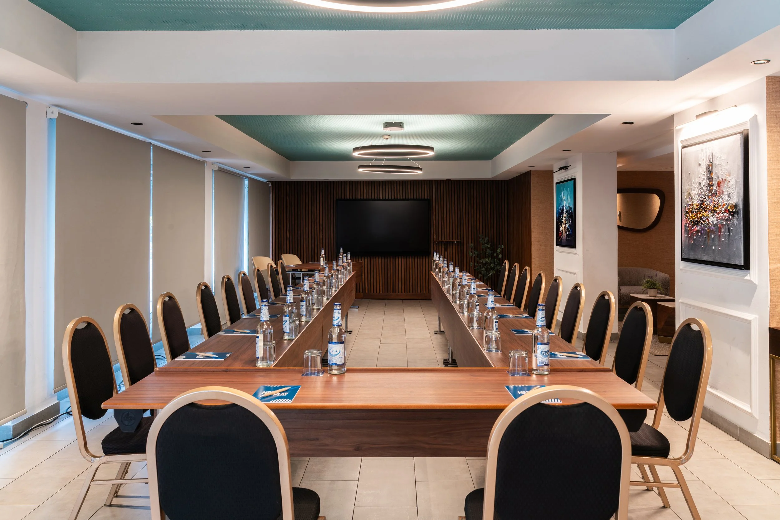 Salle de réunion en U avec tables en bois, entourée de chaises avec des bouteilles d'eau et des verres, écran TV mural au fond, œuvres d'art sur les murs, éclairage moderne suspendu.