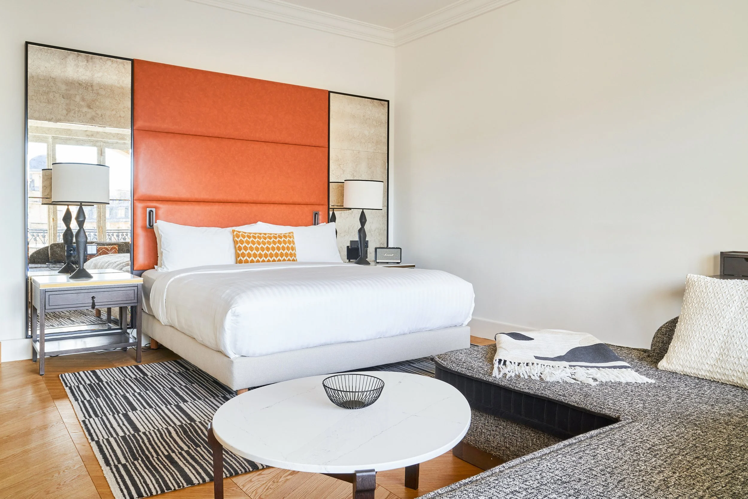 Chambre d'hôtel moderne avec un lit king-size, une tête de lit orange, deux lampes de chevet, une table basse ronde et un canapé avec des coussins et une couverture, vue sur un balcon avec des fenêtres en arrière-plan.