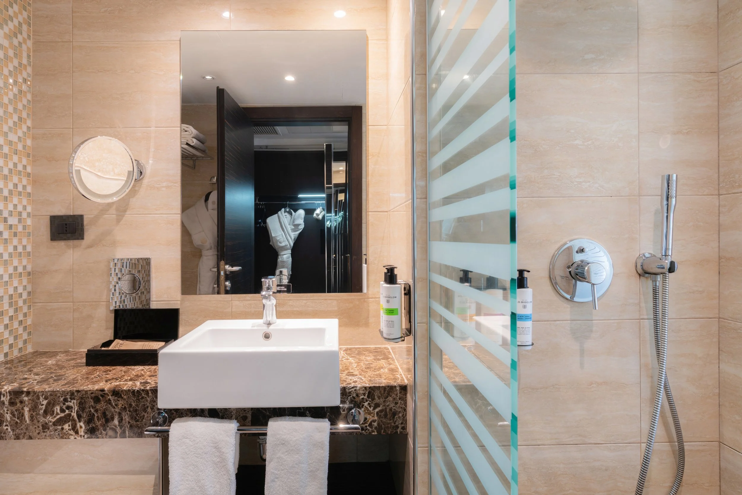 Salle de bain moderne avec un lavabo blanc sur un comptoir en marbre, un grand miroir, une armoire derrière la porte, et une cabine de douche avec paroi en verre.