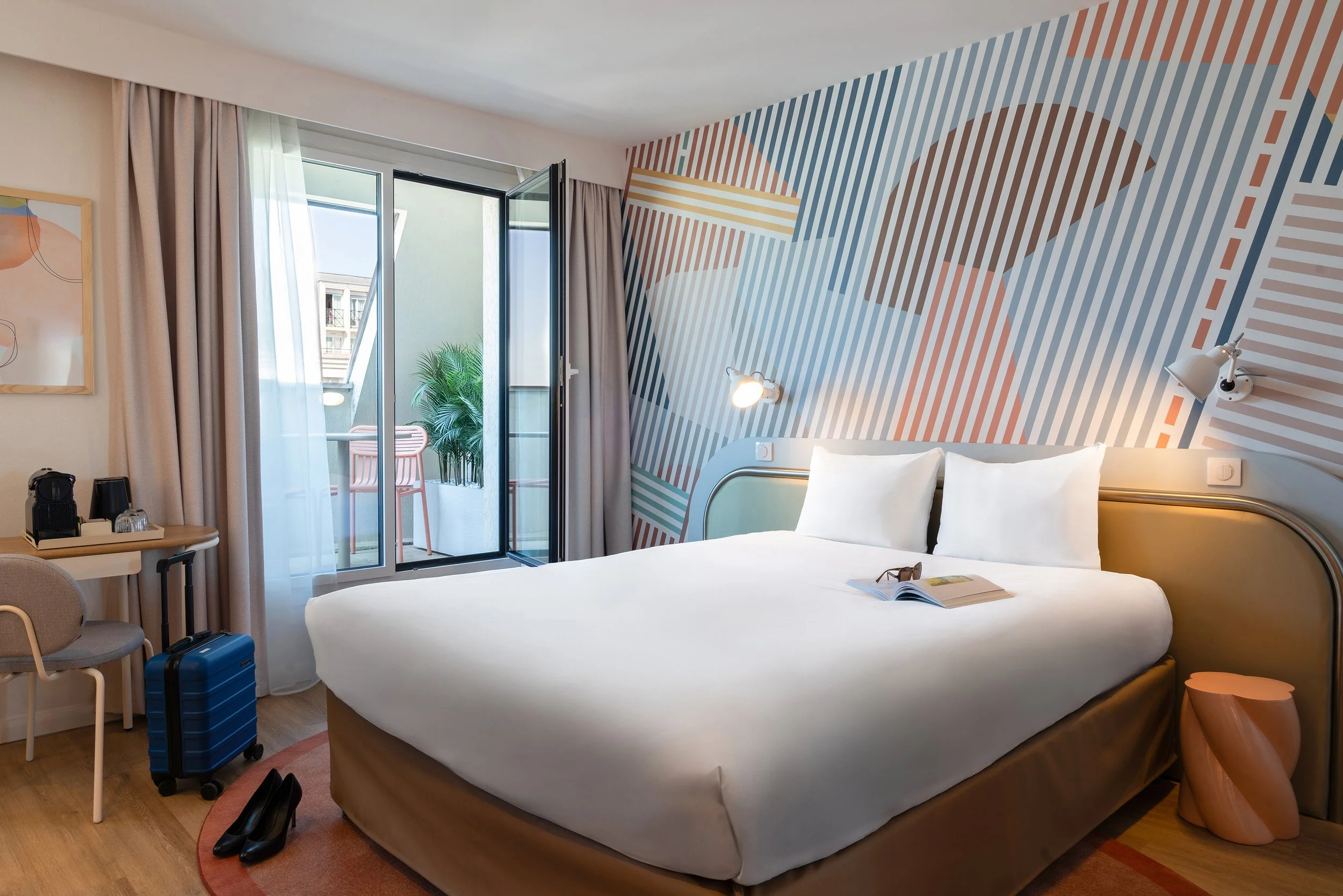 Chambre d'hôtel moderne avec un lit, une table avec un café, une valise, un décor mural géométrique, un balcon avec une plante, une paire de chaussures et une paire de lunettes