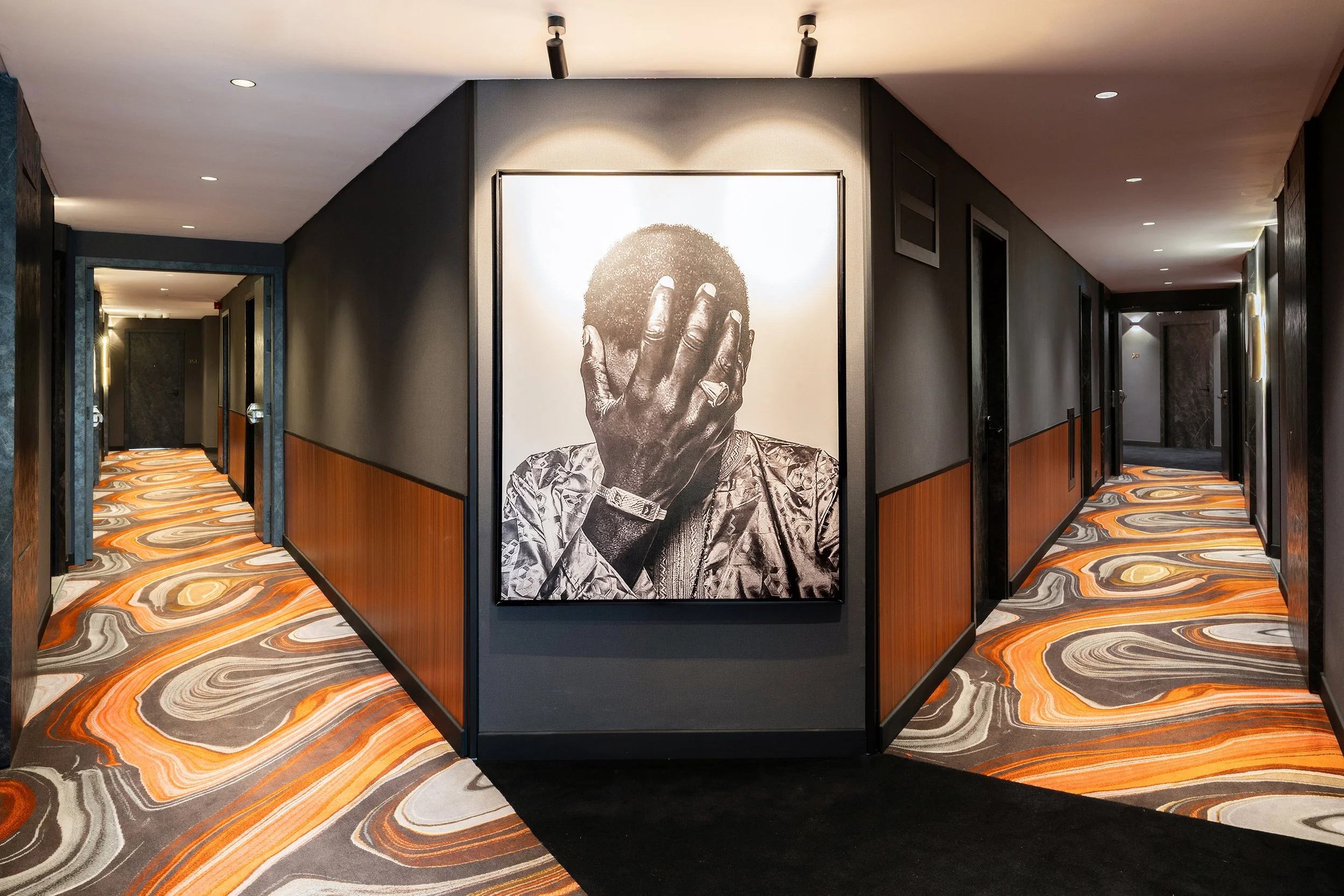 Couloir d'hôtel avec mur noir, décor en bois, tapis à motifs orange, gris, marron et beige, et grande photo en noir et blanc d'une personne avec la main sur le visage.