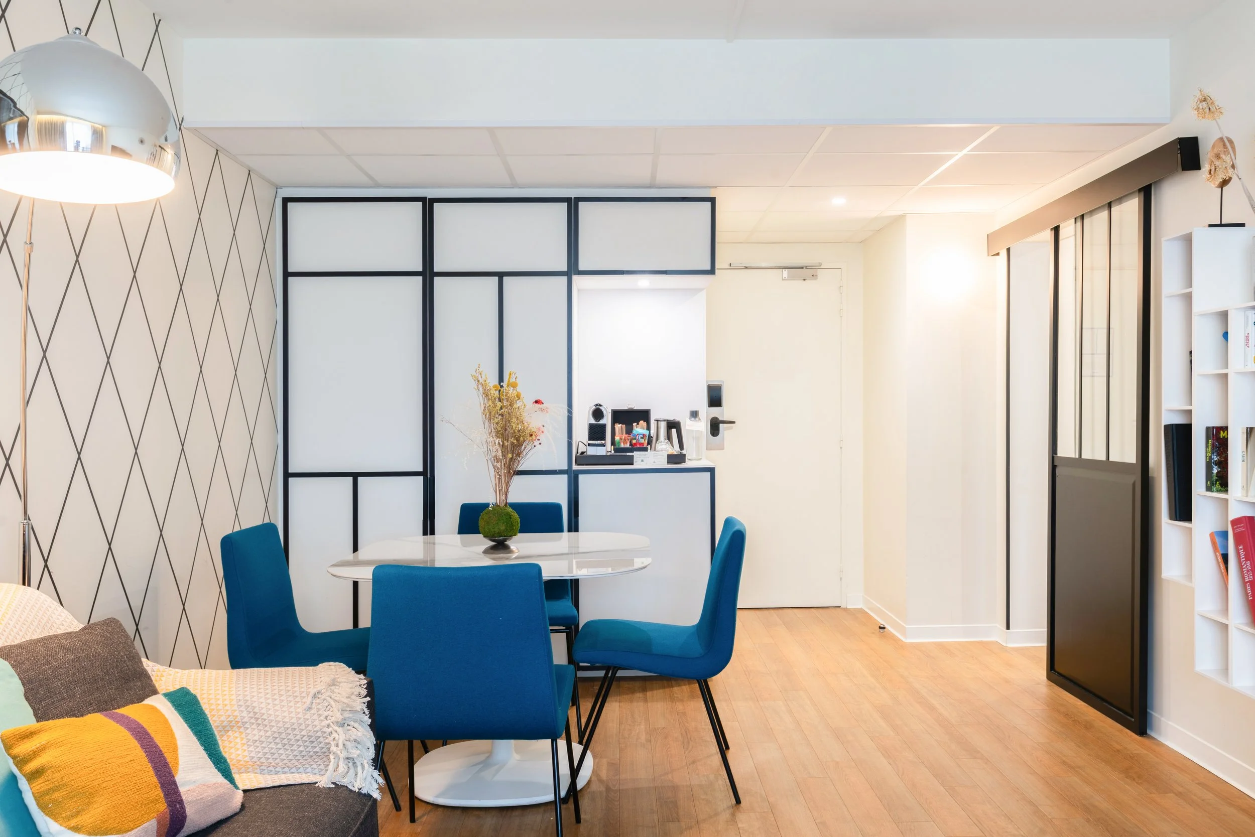 Salon avec canapé gris, coussins colorés, fauteuils bleus autour d'une table ronde avec vase floral, mur en papier peint à losanges, coin bar avec machine à café, étagère blanche avec livres et téléviseur noir, sol en bois clair.