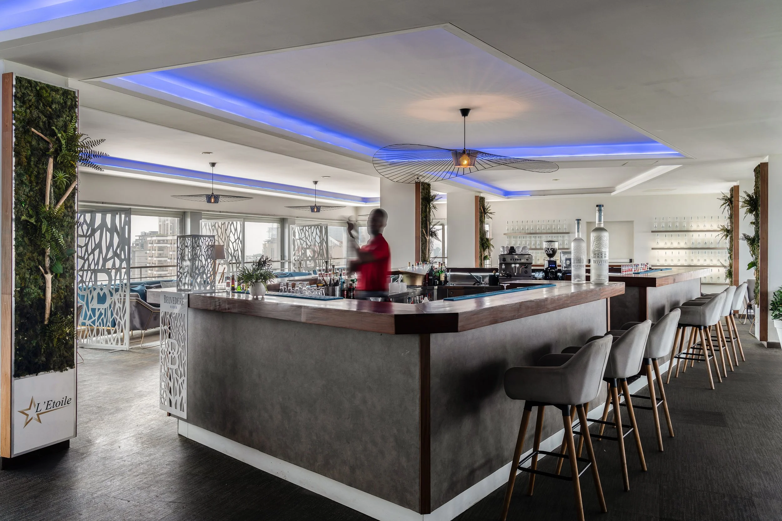 Bar à cocktail moderne dans un hôtel avec comptoir en bois, chaises en tissu gris, plantes vertes, et ambiance lumineuse avec accents bleus au plafond.