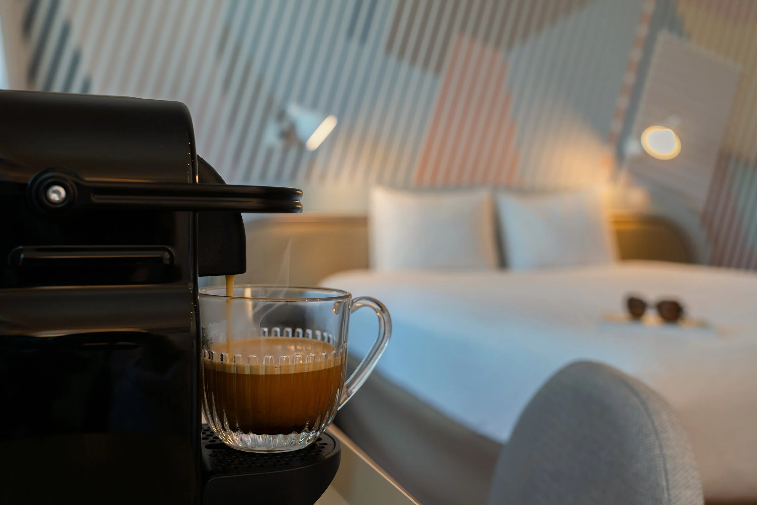 Une machine à café verse du café chaud dans une tasse en verre. En arrière-plan, un lit avec des oreillers dans une chambre moderne, avec des lunettes de soleil posées sur la couverture.