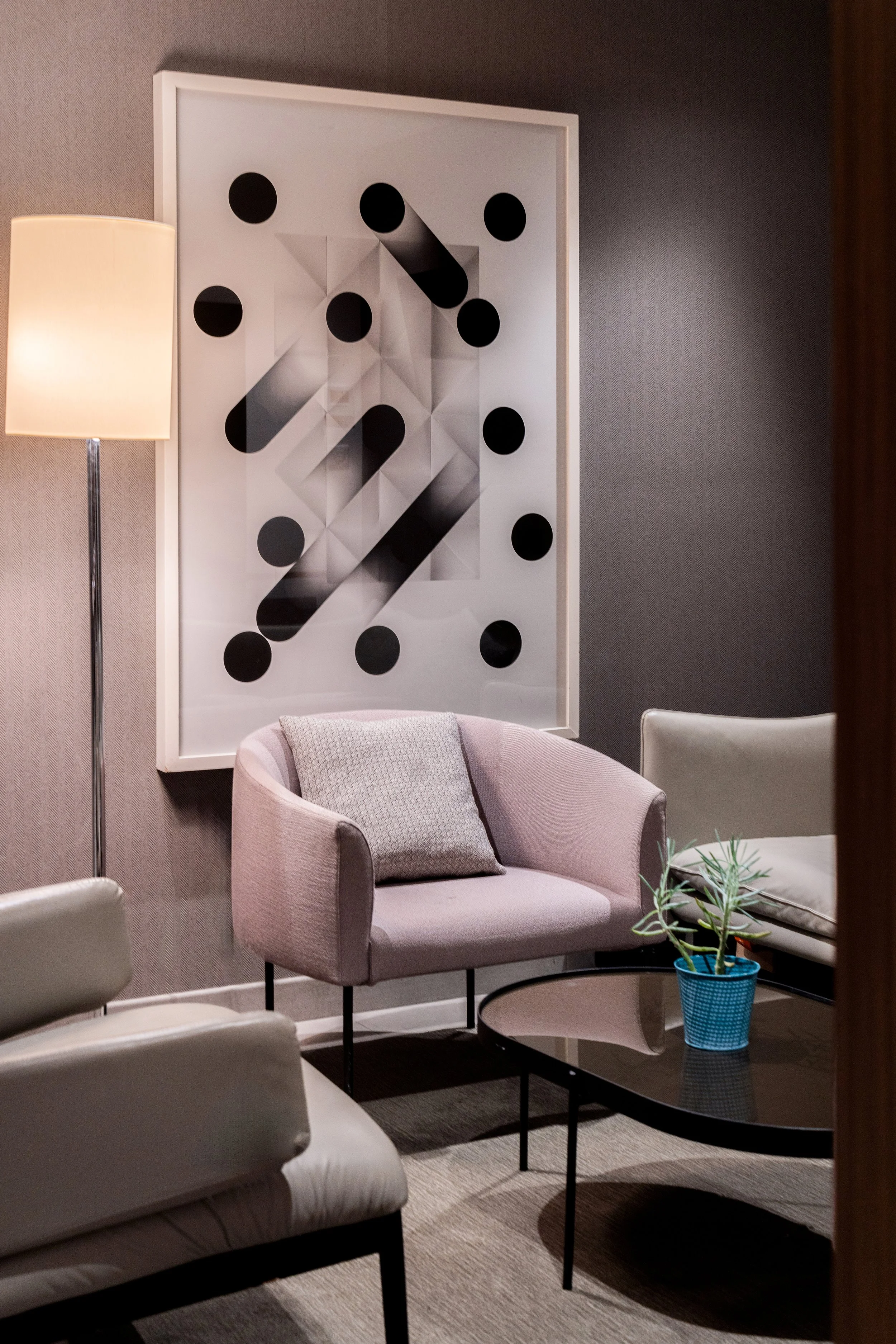 Coin salon avec une lampe, un fauteuil beige avec un coussin, une table basse noire avec une plante en pot bleu, et une œuvre d'art abstraite avec des cercles noirs et des formes géométriques au mur.
