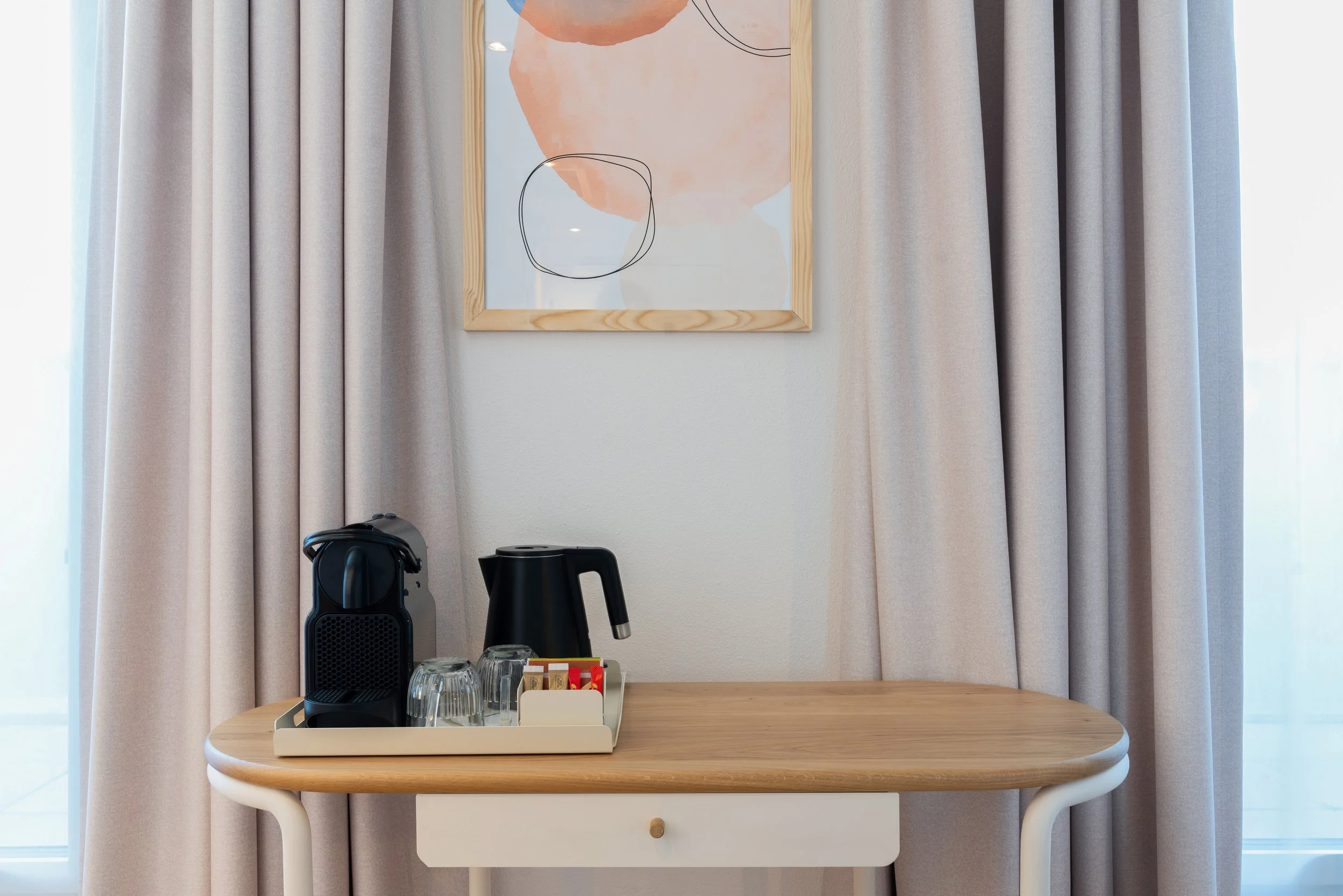 Table en bois avec une machine à café, une bouilloire électrique, quelques tasses et sachets de café, devant des rideaux beige clair et un tableau abstrait sur le mur.