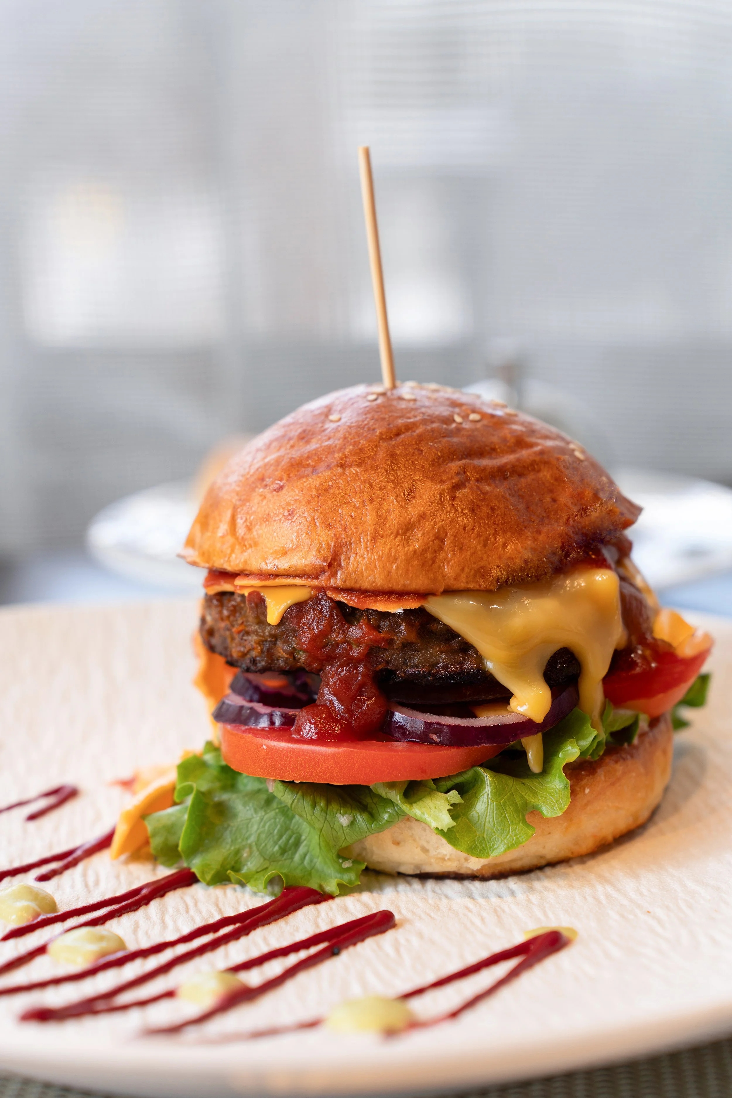 Un hamburger composé d'un bun croustillant, un steak haché, fromage fondu, tranches de tomate, oignons rouges, laitue, et ketchup, décoré avec des sauces sur une assiette