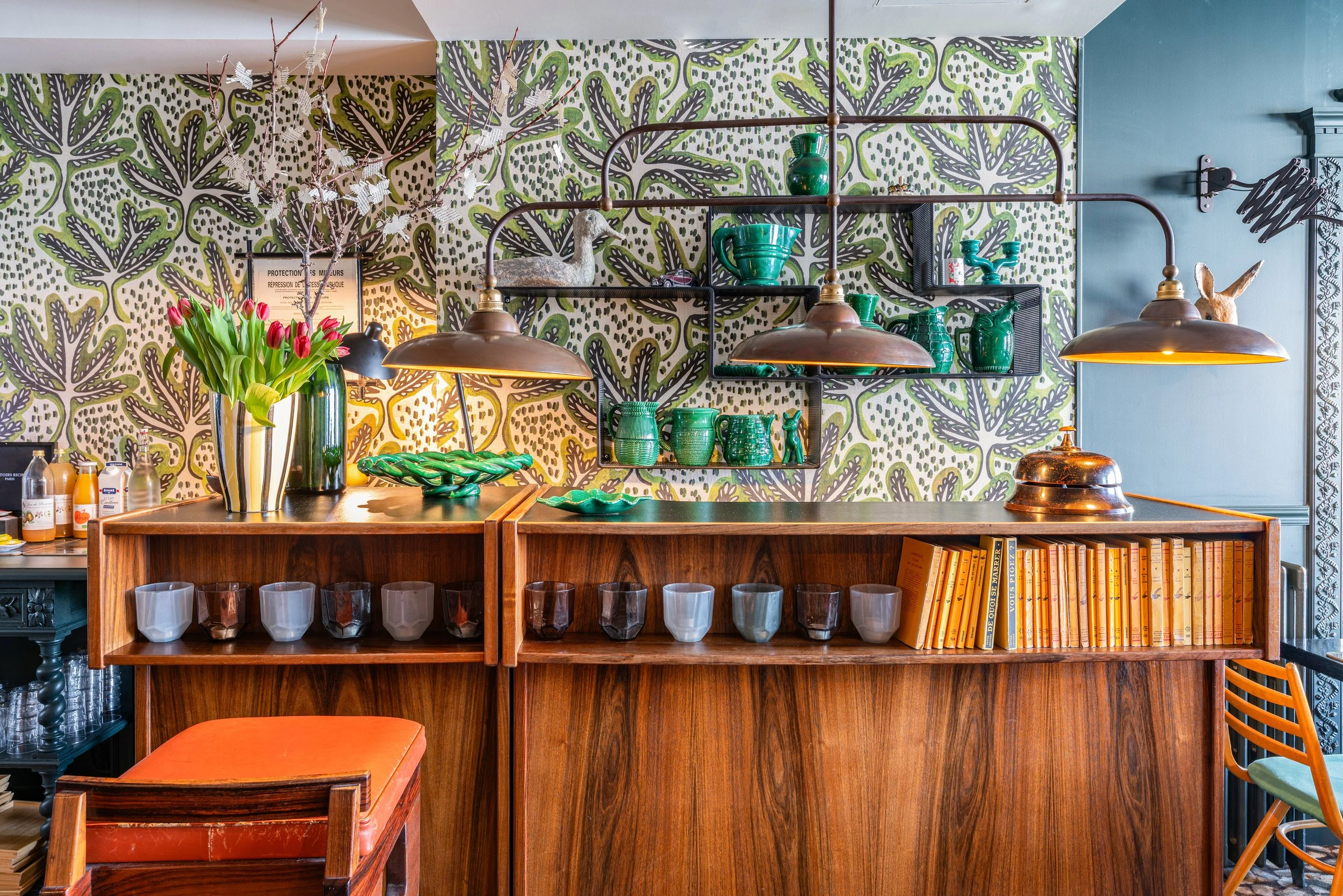 Intérieur coloré d'un restaurant avec un comptoir en bois, étagères décoratives contenant des vases, des tasses vertes, des livres jaunes, décorations en porcelaine, un vase avec des tulipes, et un fond mural avec un motif de feuilles vertes et viole