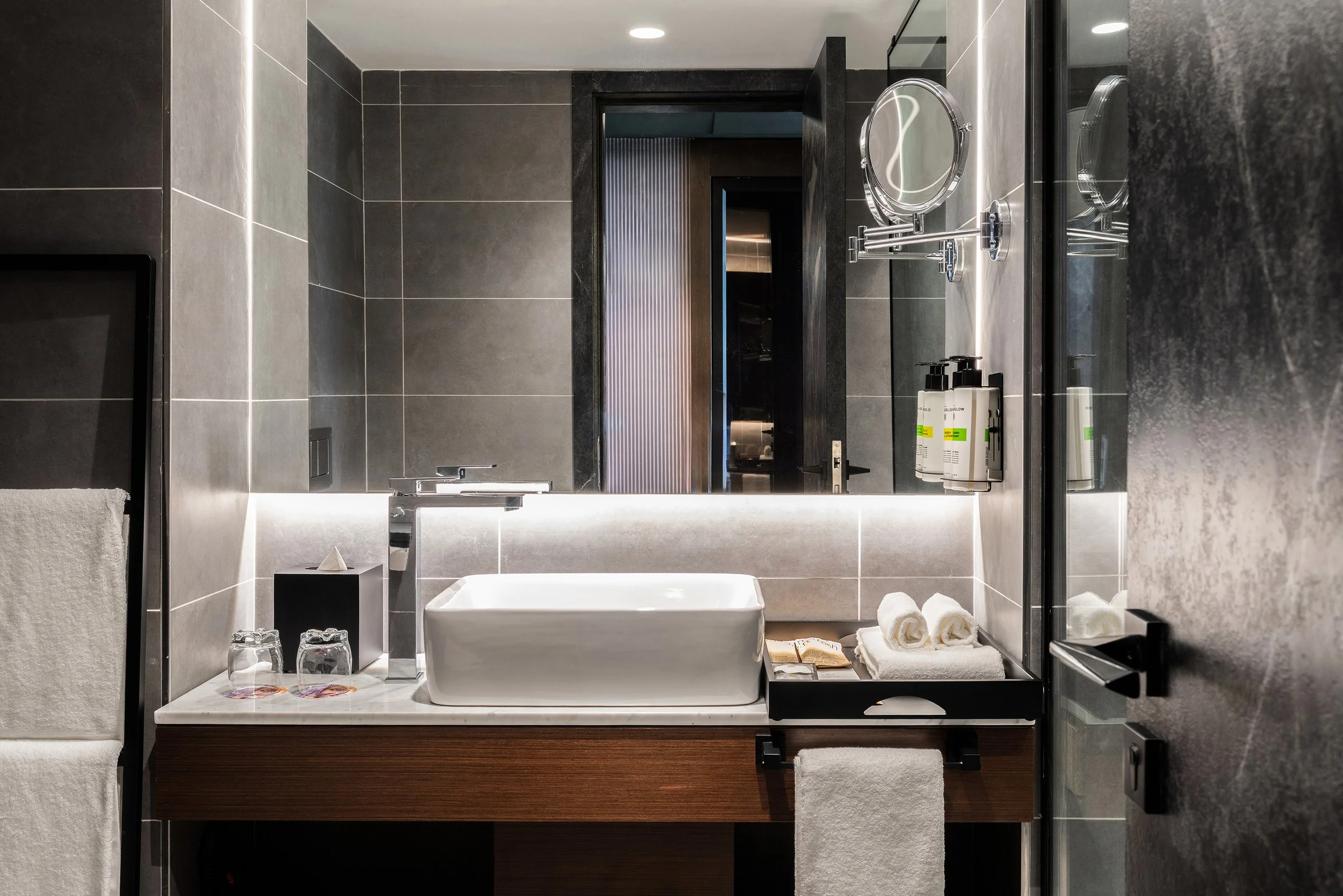 Salle de bain moderne avec un lavabo blanc carré, miroir rond, serviettes blanches, produits de toilette, murs gris foncé, éclairage intégré.
