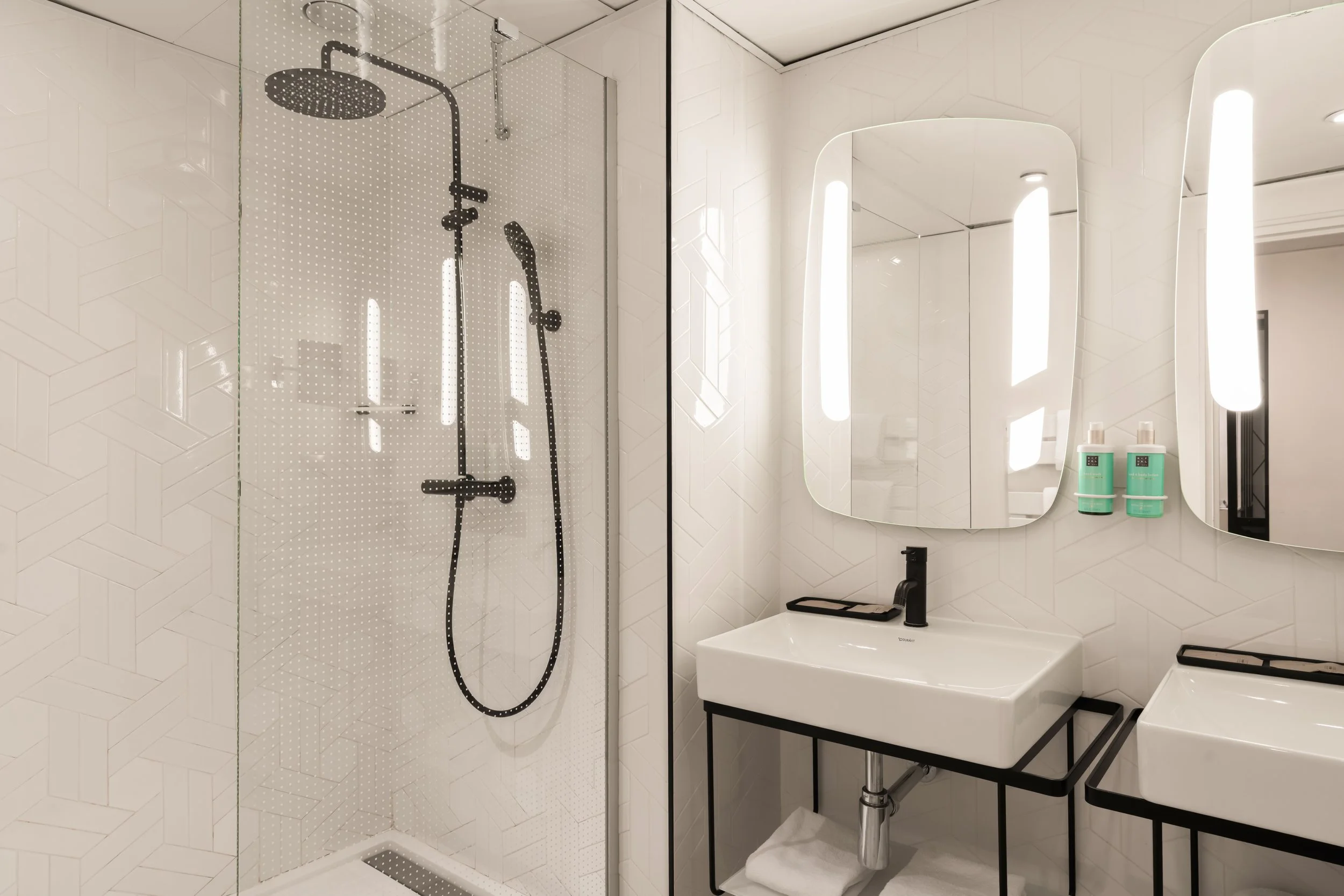 Salle de bain moderne avec douche à l'italienne, miroir(s) avec éclairage intégré, lavabo à cadre métallique, et deux bouteilles de produits de lavage