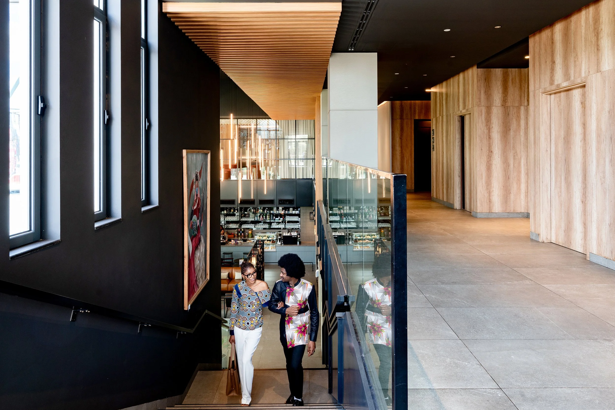 Un couple discute en montant les escaliers dans un hall moderne et élégant, avec un bar en arrière-plan et un décor en bois et en noir.