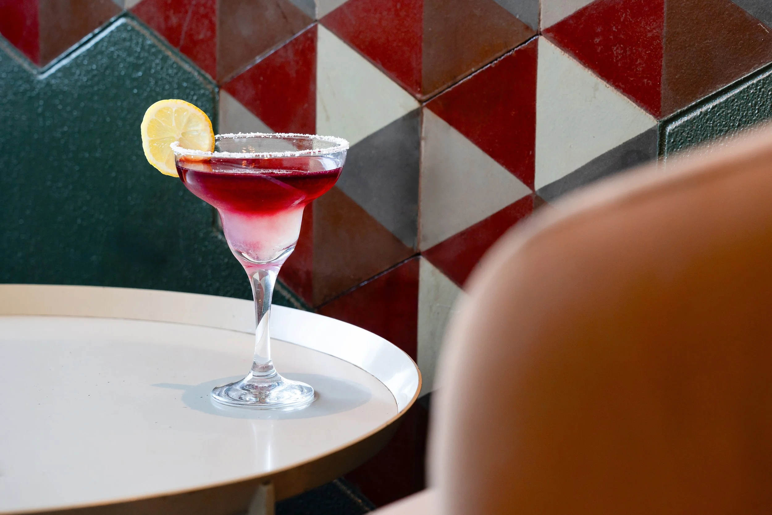 Un verre à cocktail rouge avec une tranche de citron, placé sur une table blanche, avec un fond de mosaïque colorée.