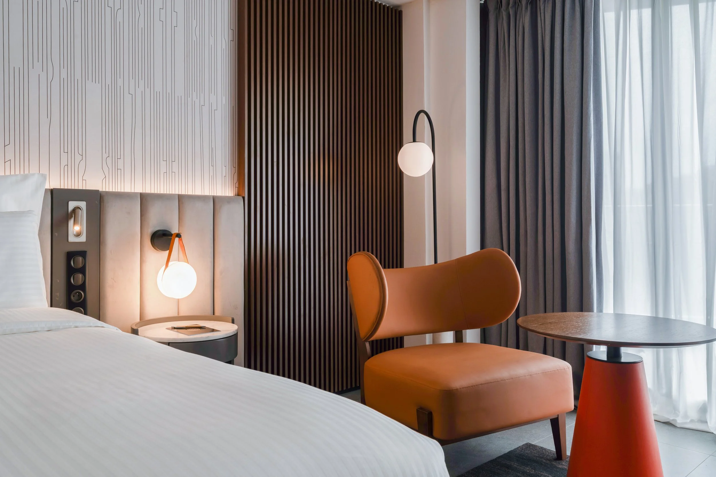 Chambre moderne avec lit, tables de nuit, lampes suspendues, fauteuil orange et rideaux sur la fenêtre.