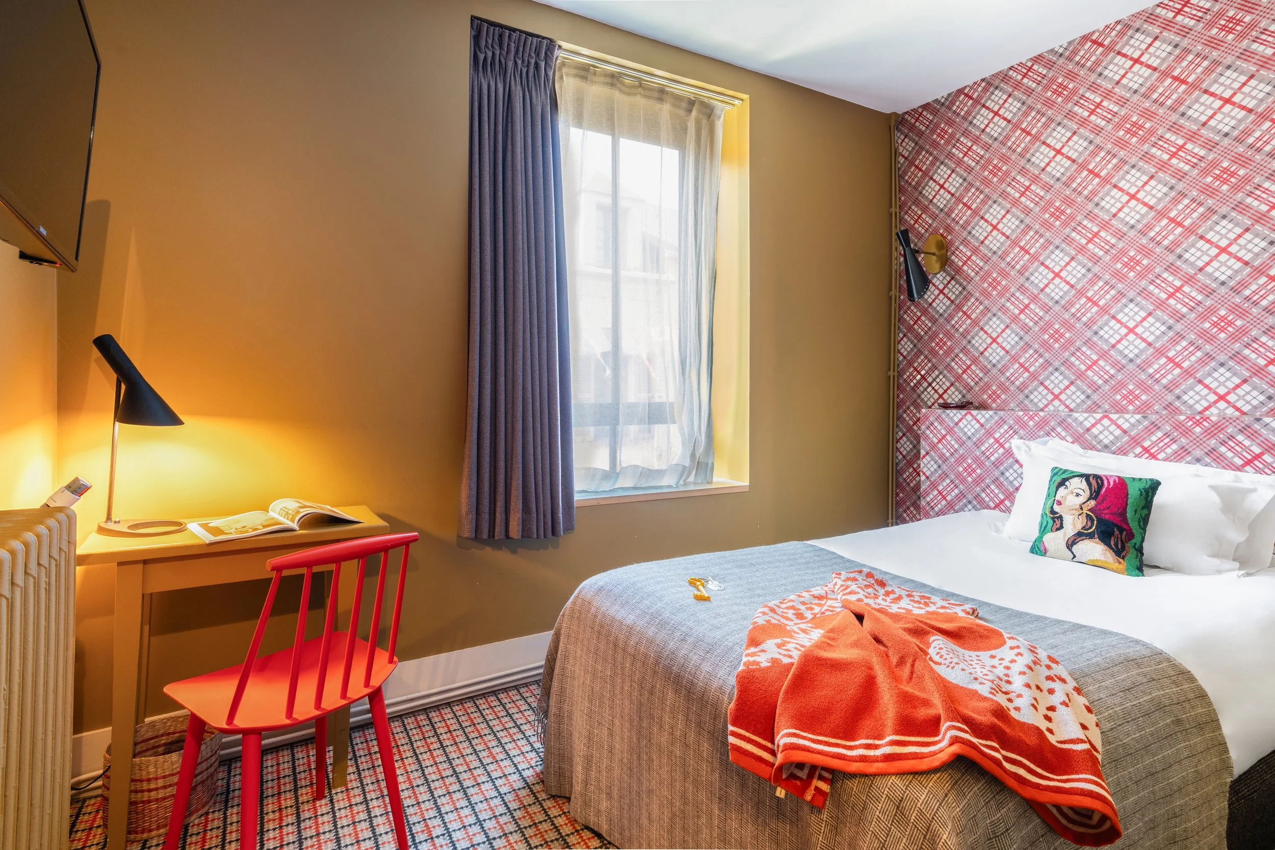 Chambre d'hôtel avec un lit, un bureau avec une lampe et un ordinateur, une fenêtre avec des rideaux, et un mur avec un motif en tartan rouge et blanc.