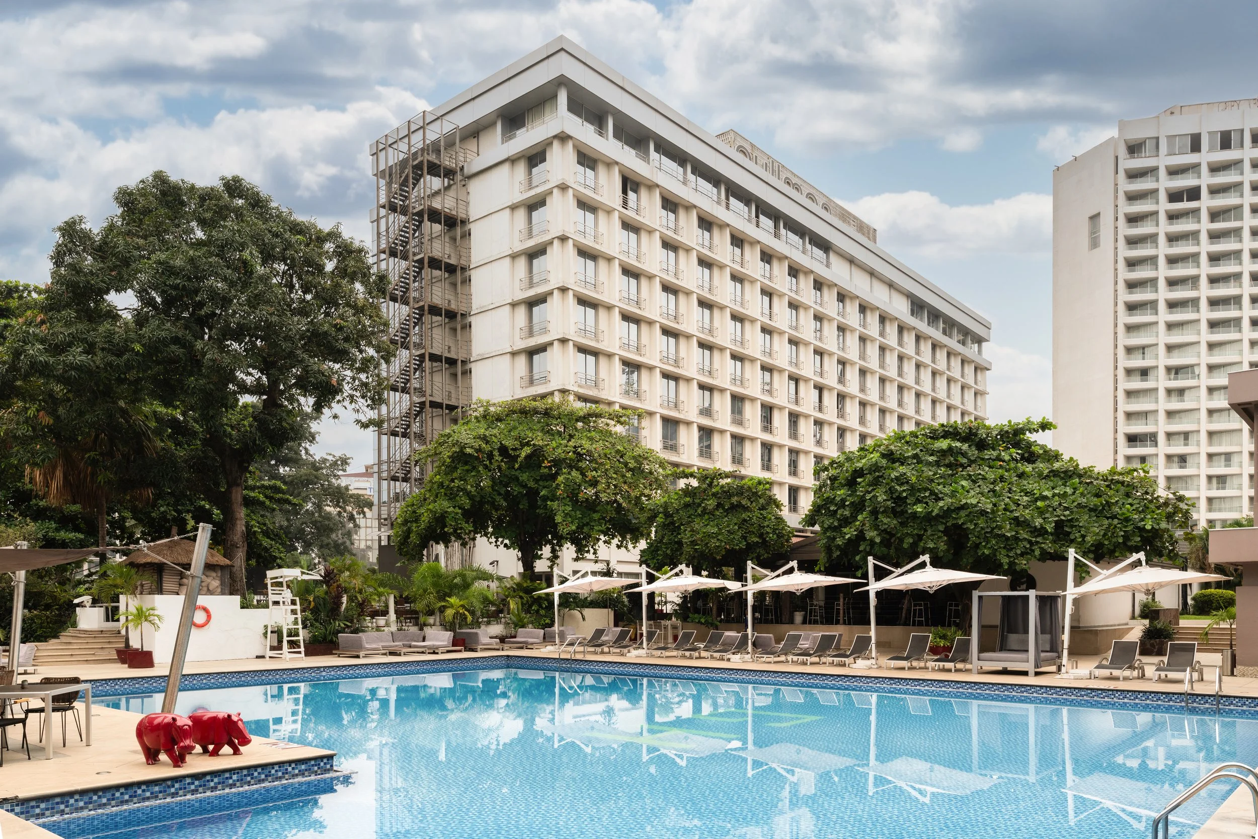 Piscine extérieure dans un complexe hôtelier avec chaises longues, parasols blancs, arbres verts et grands immeubles blancs en arrière-plan, sous un ciel partiellement nuageux.