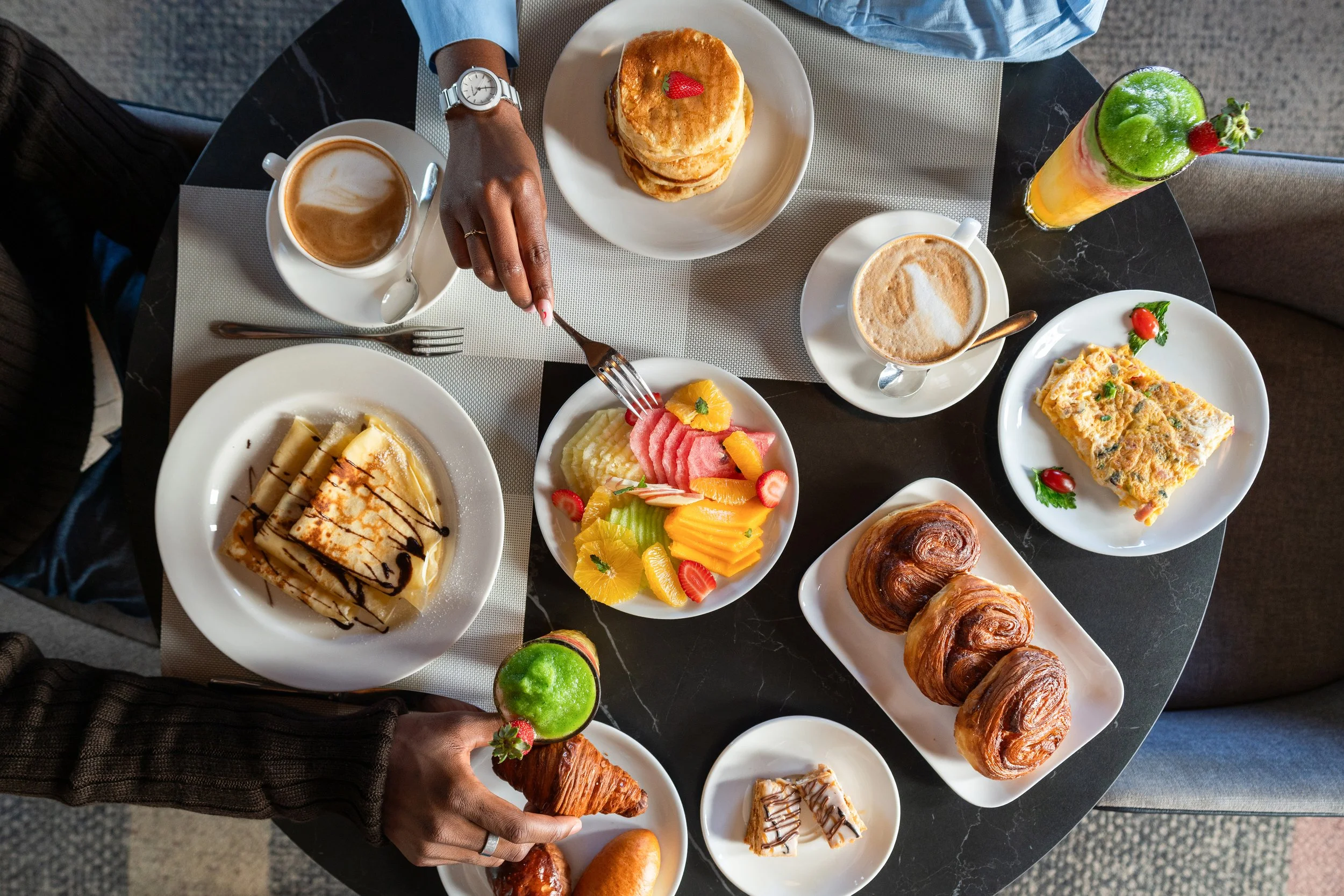 Petit déjeuner varié avec des crêpes, quiche, croissants, gâteau, fruits, café, latte, thé et boissons fruitées sur une table ronde