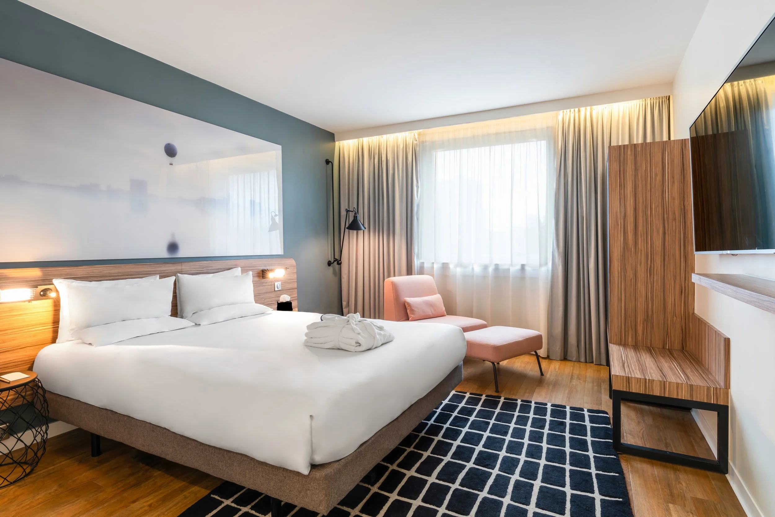 Chambre d'hôtel moderne avec un lit, une chaise rose, un meuble en bois, une télévision, des rideaux clairs et un tapis noir et blanc.