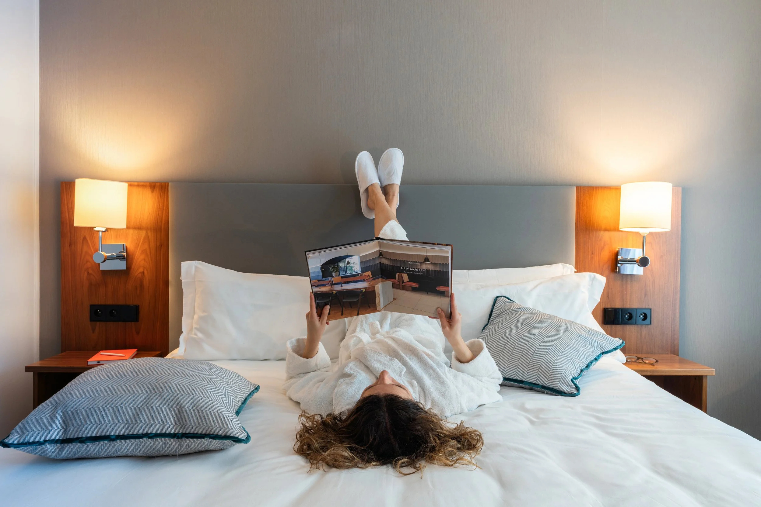 Femme allongée dans un lit, lisant un magazine, deux lampes allumées de chaque côté du lit.