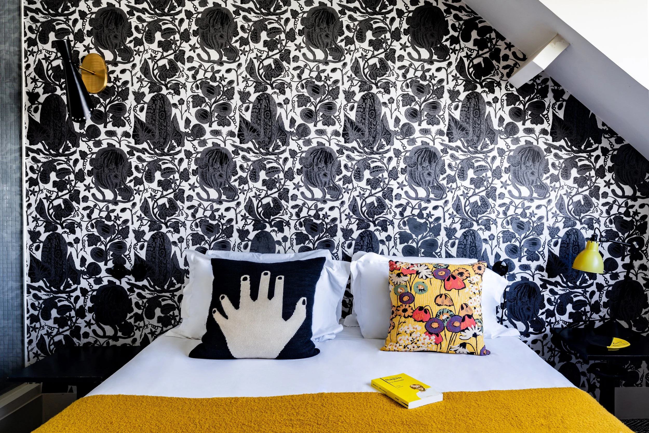 Chambre avec papier peint noir et blanc à motif floral, deux oreillers décoratifs, un avec main stylisée et l'autre avec fleurs colorées, une lampe jaune et une couverture jaune sur le lit.