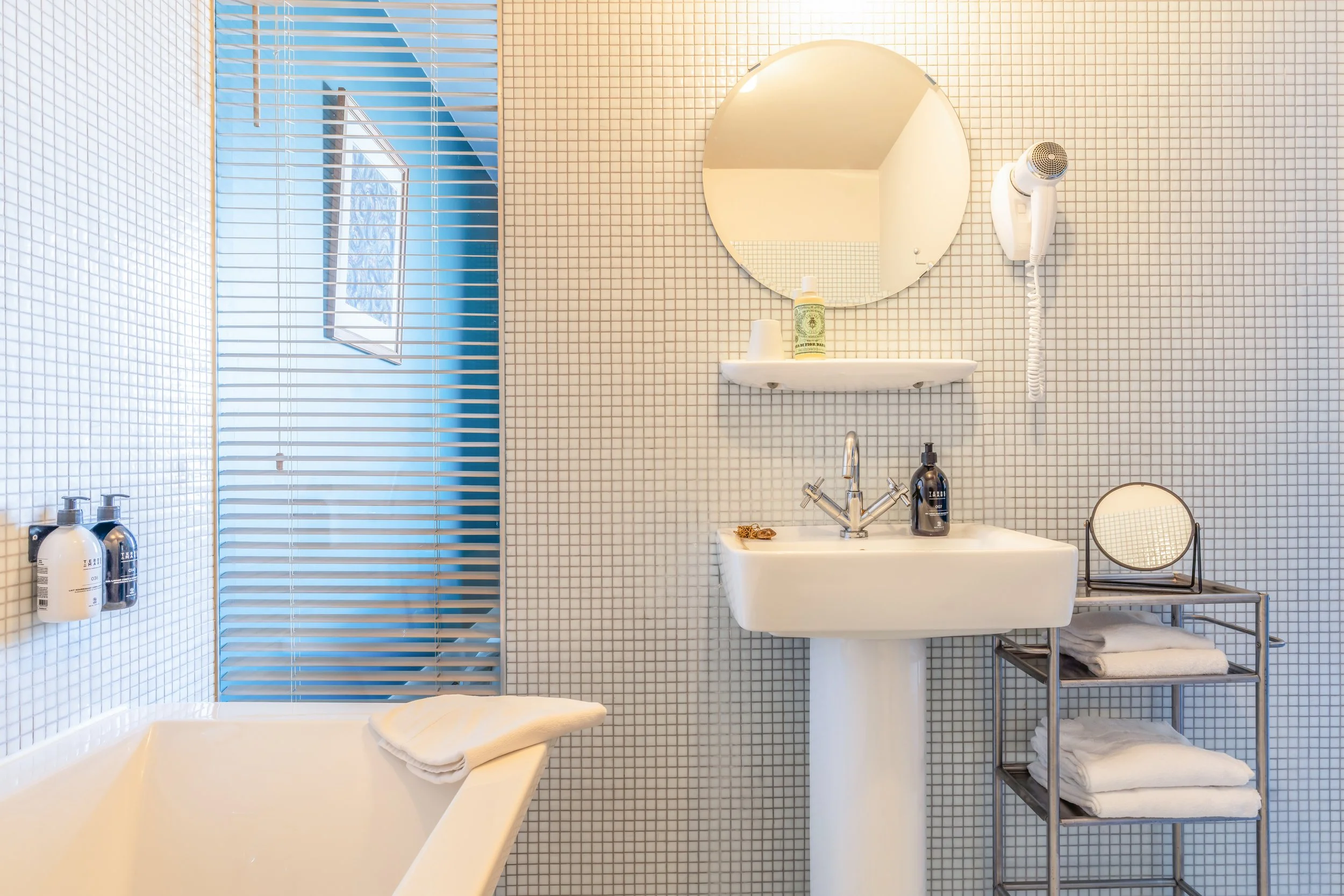 Salle de bain moderne avec baignoire, lavabo, miroir rond, sèche-cheveux et étagère avec serviettes et produits de toilette.