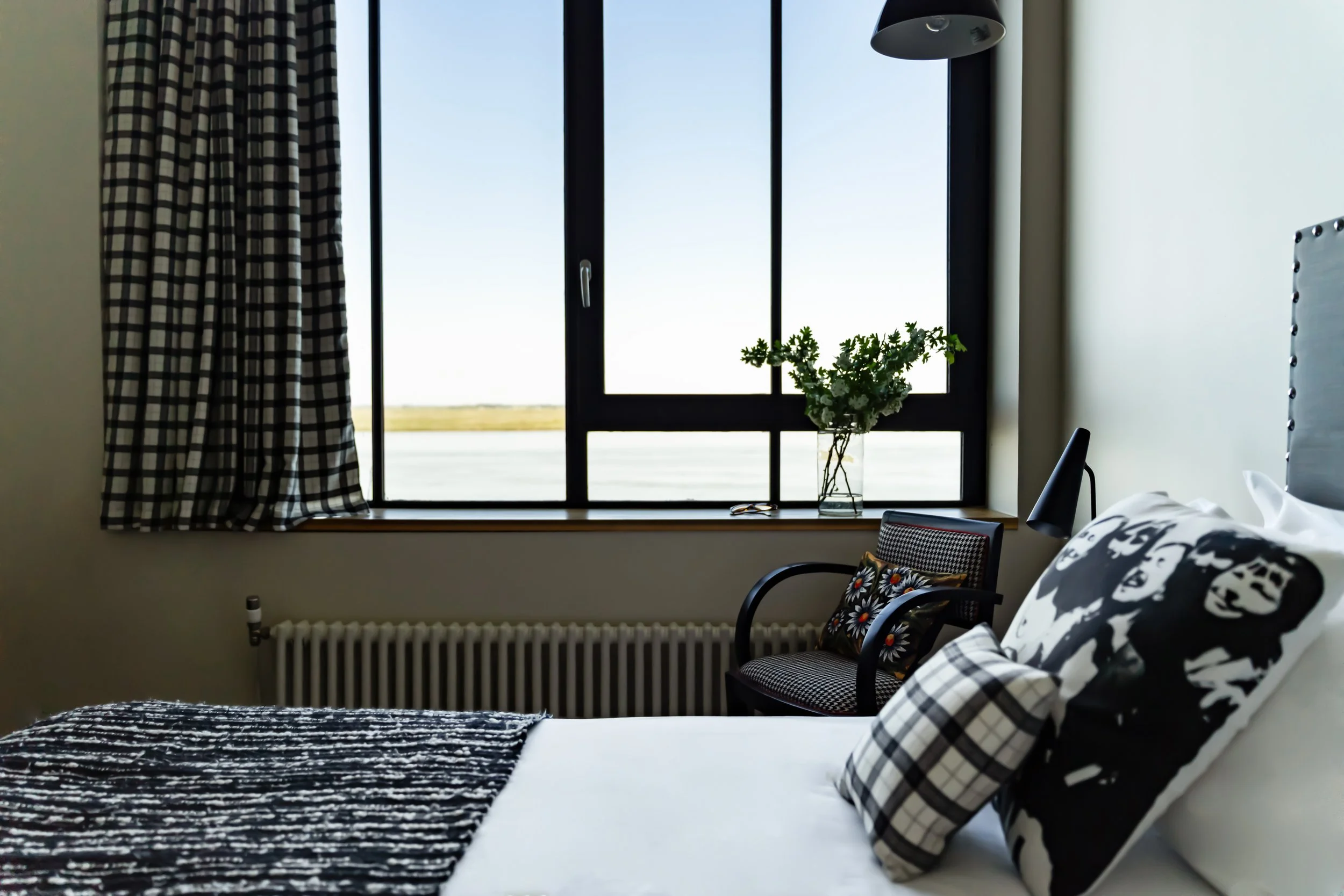 Chambre avec un lit, une fenêtre grande laissant entrer de la lumière naturelle, rideaux à carreaux, une plante dans un vase sur le rebord de la fenêtre, une chaise avec un coussin décoratif, un lampadaire noir, et un radiateur sous la fenêtre.